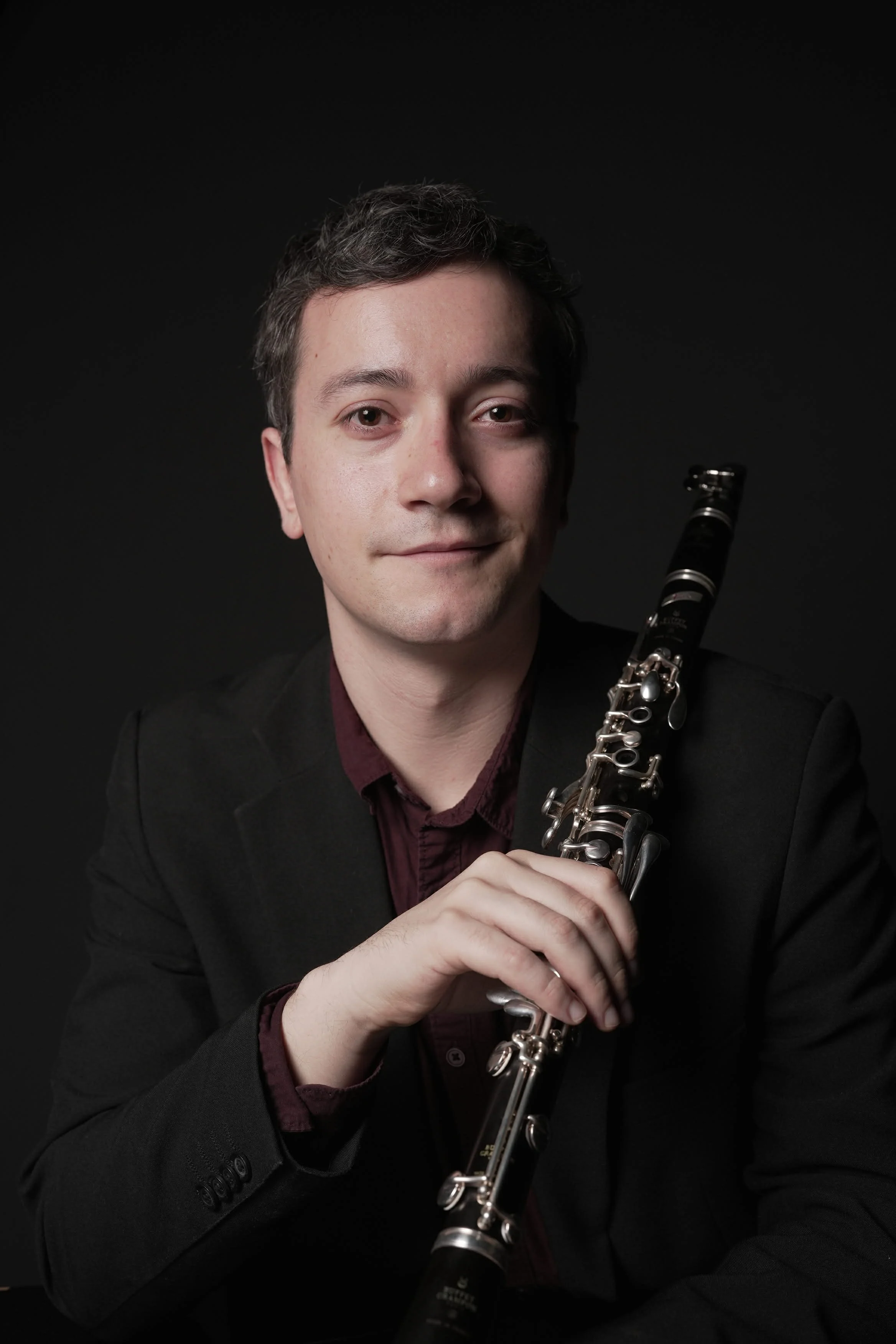 Portrait of a young man in a black suit holding a clarinet against a black background.