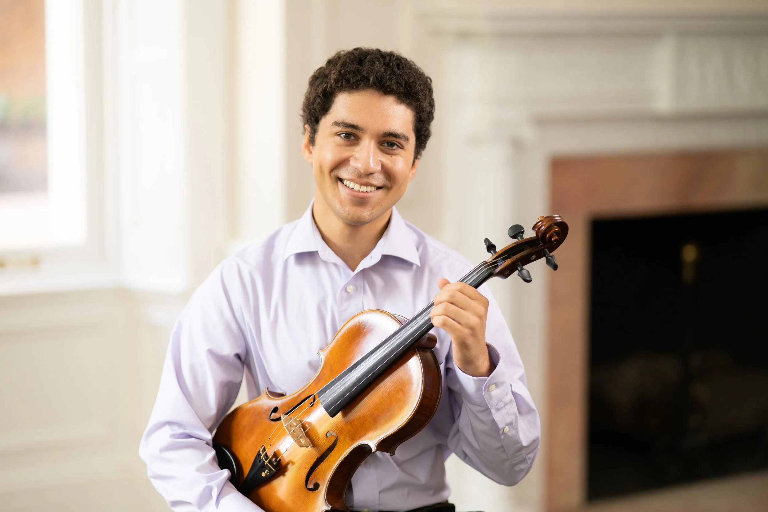 A smiling man holding a violin indoors near a window and fireplace.
