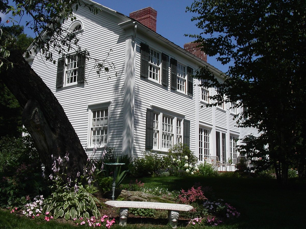 A white two-story house with black shutters, surrounded by a lush garden with flowers and trees, under a clear blue sky.