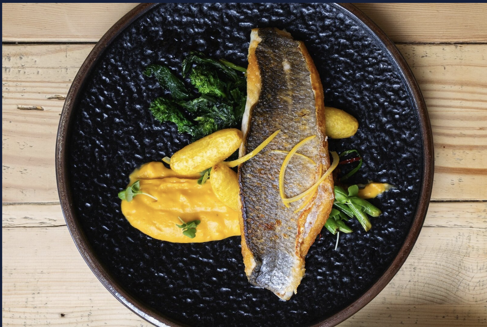 A plate of cooked fish with crispy skin, accompanied by yellow sauce, baby potatoes, sautéed greens, and garnished with microgreens, served on a black textured plate on a wooden surface.