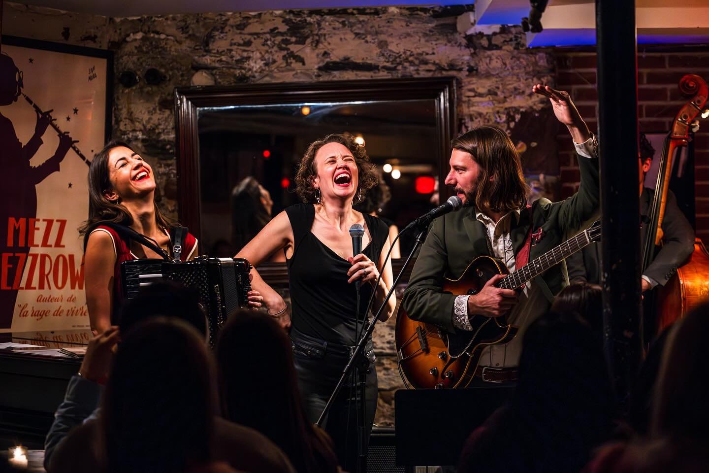 Three musicians performing on stage, singing and playing instruments, with a crowd in front. The woman on the left is playing an accordion and smiling, the woman in the middle is holding a microphone and laughing, and the man on the right is playing a guitar and raising his hand.