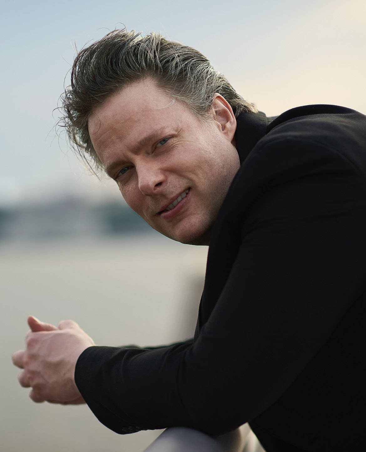 A man with light-colored hair and a slight smile, wearing a black jacket, leaning on a railing outdoors, with water and a cloudy sky in the background.