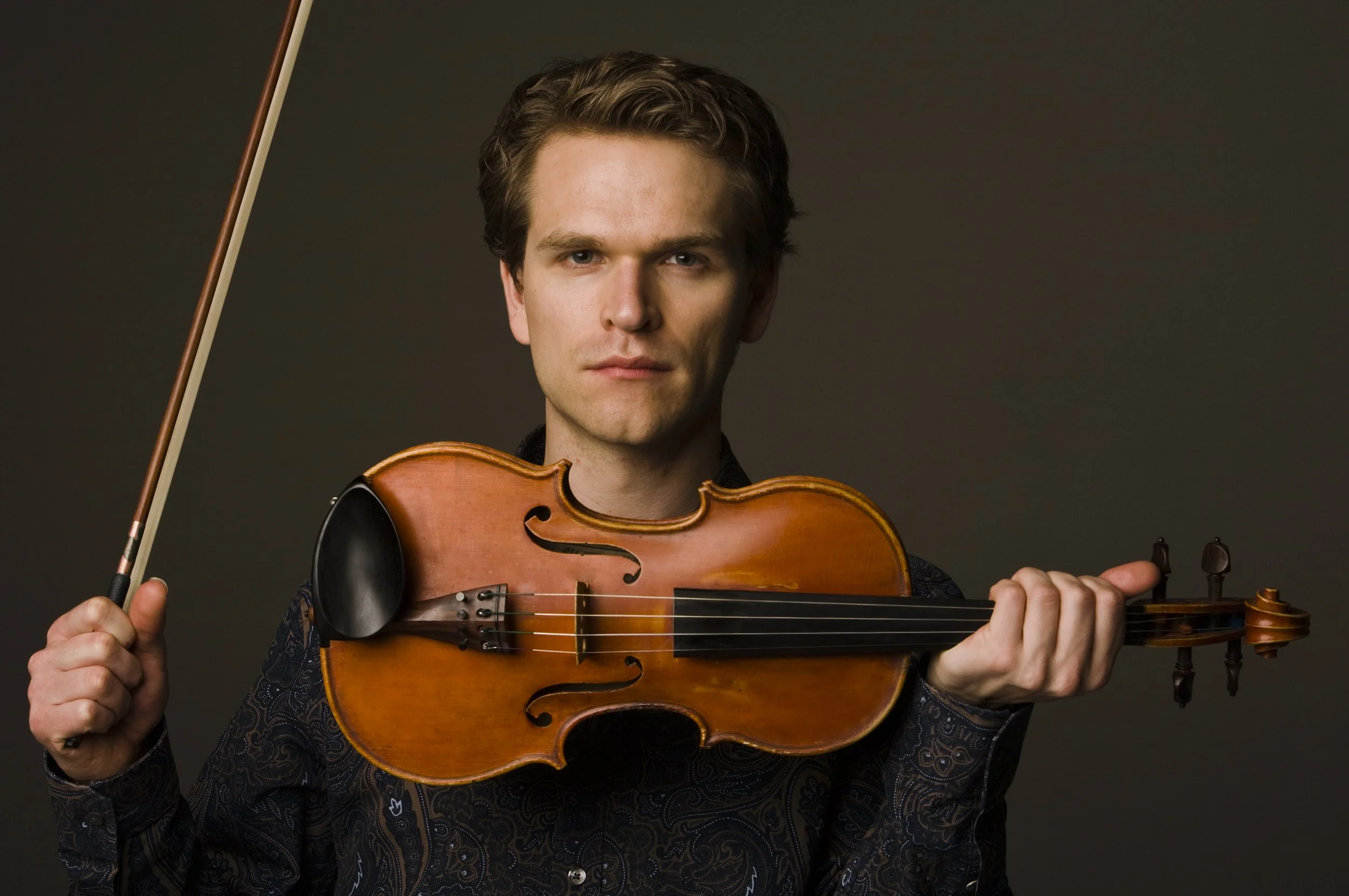 A man with dark hair holding a violin under his chin and a bow in his right hand, looking directly at the camera against a dark background.