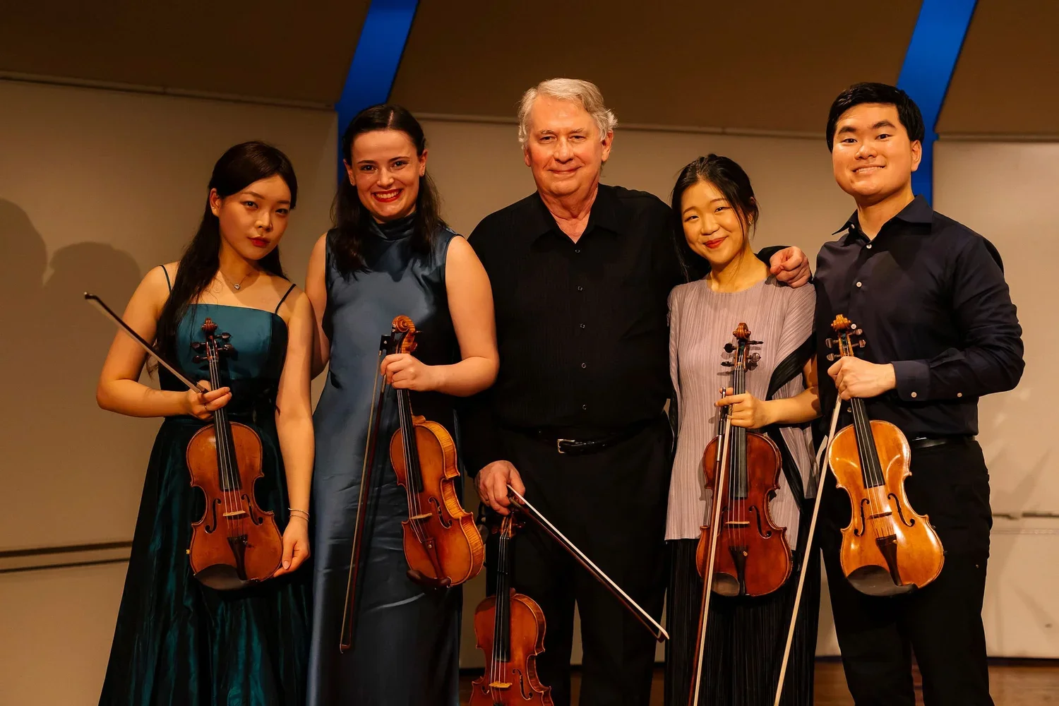 Five people, including four violinists and their conductor, posing with their violins on stage after a performance.
