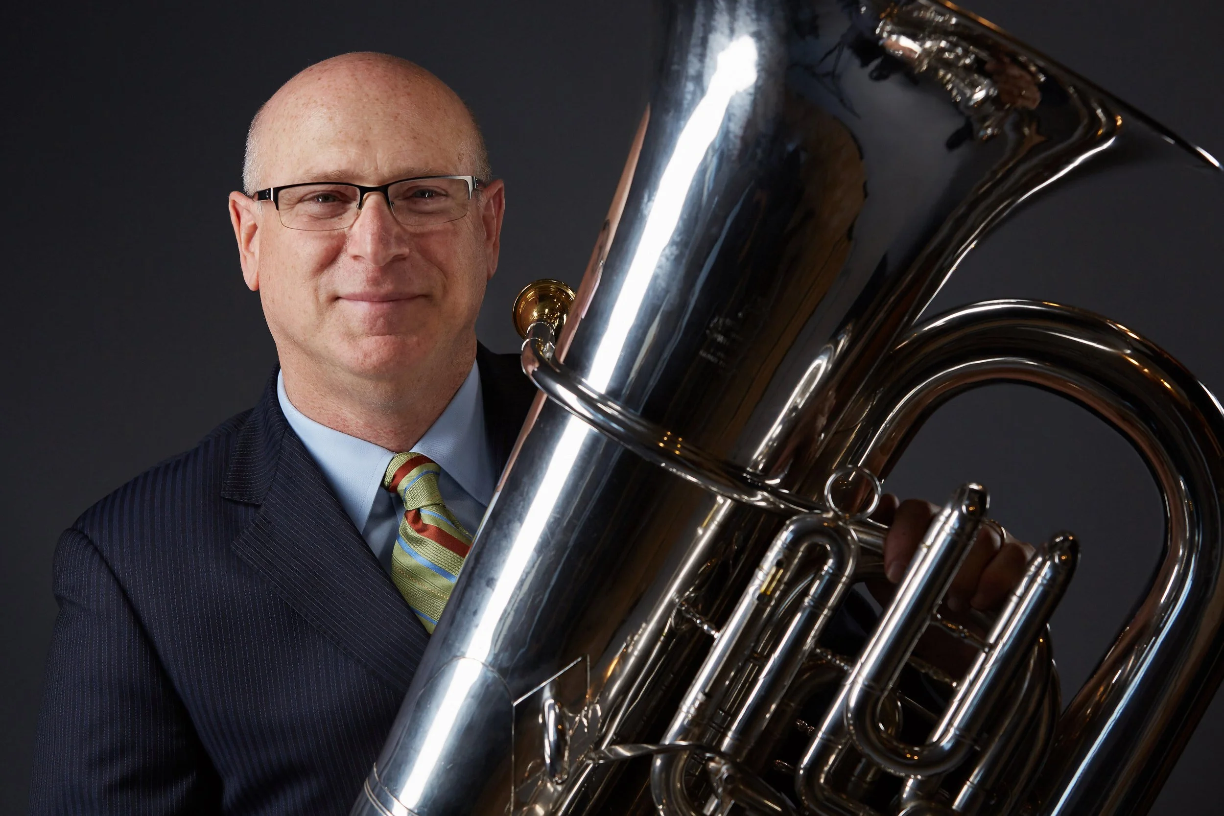 A middle-aged man with glasses in a suit and colorful striped tie holding a large, shiny brass tuba.