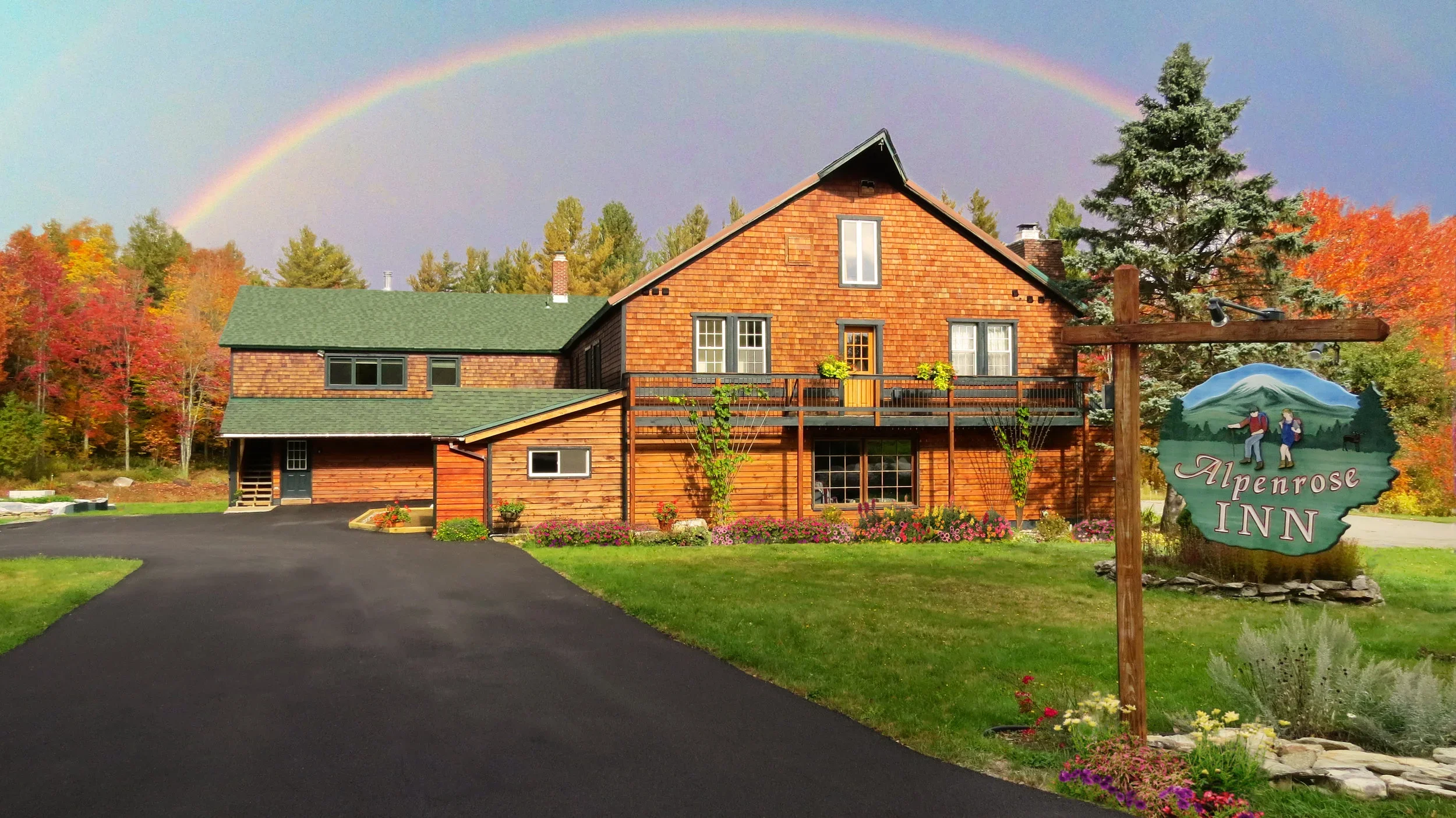A wooden lodge with a green roof, surrounded by fall foliage and flowers, under a rainbow in the sky, with a sign that reads 'Alpenrose Inn' in front.