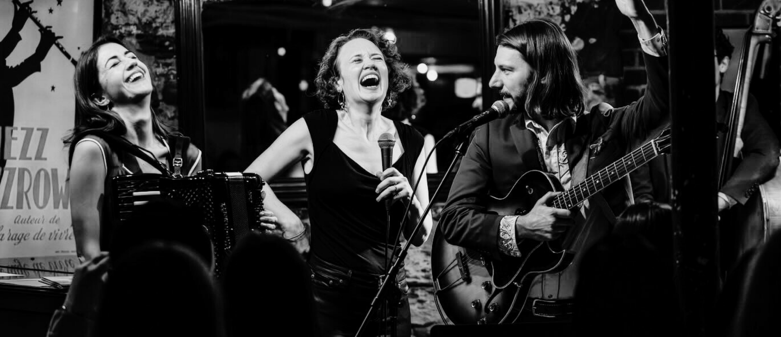 Three musicians performing on stage, one woman with an accordion, another woman singing into a microphone, and a man playing an acoustic guitar, all smiling and enjoying the moment, in a lively indoor setting.