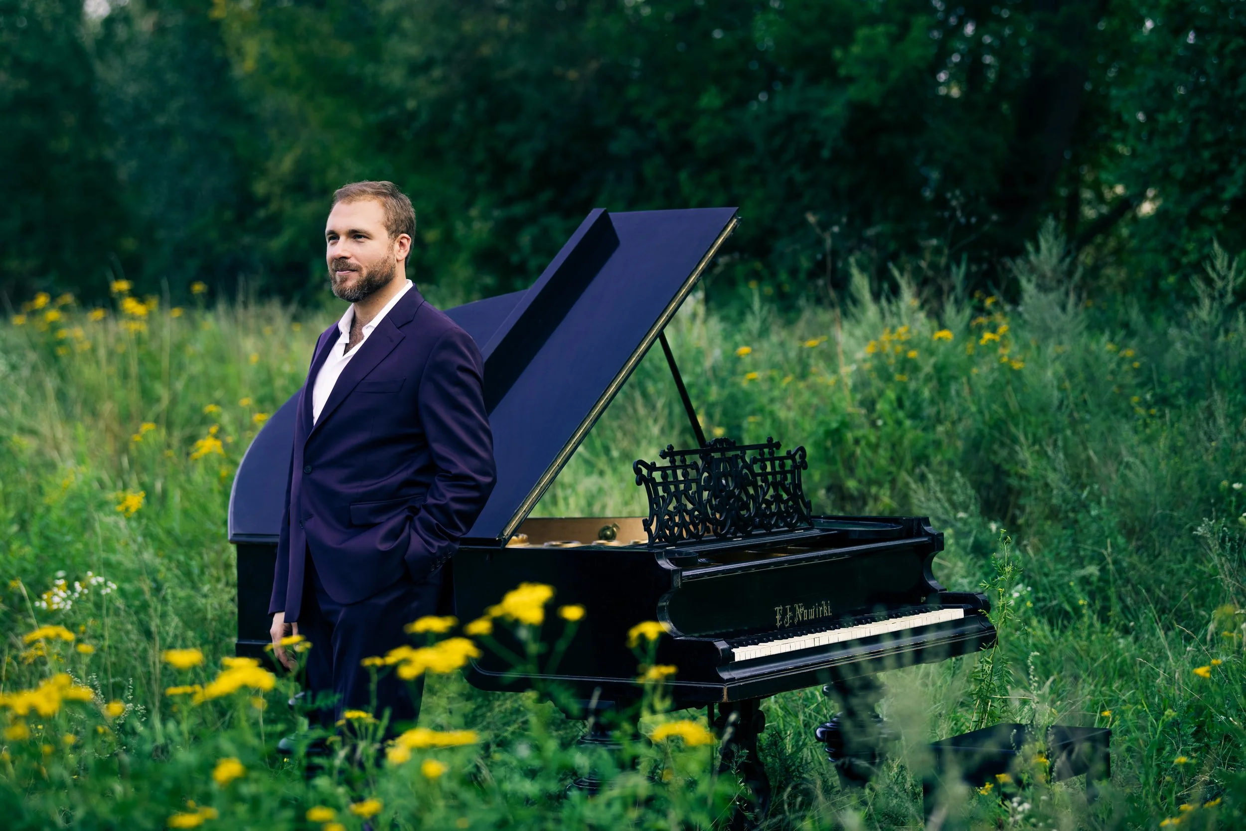 A man in a suit stands next to a black grand piano in a field with yellow flowers and green trees in the background.