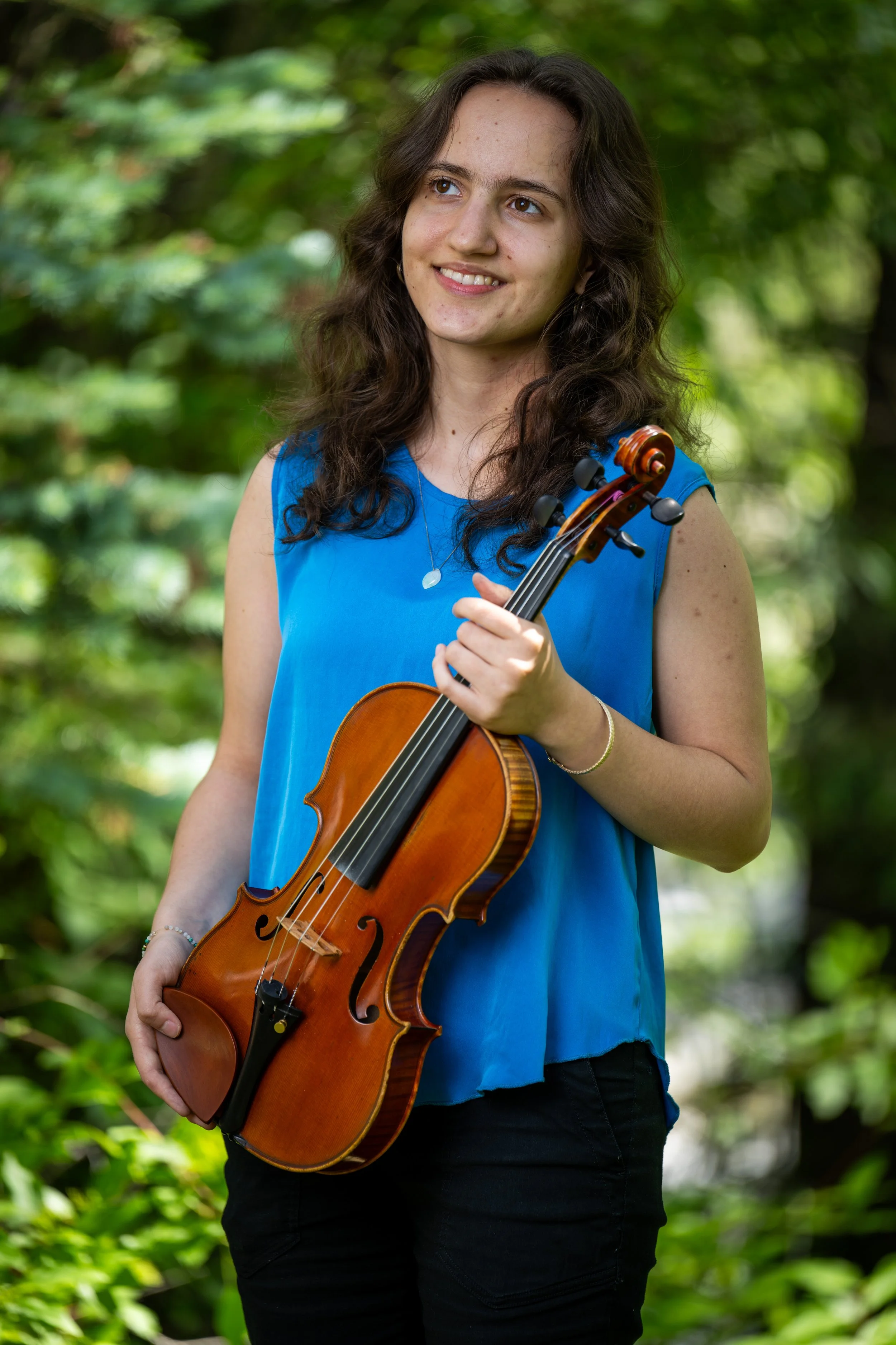 Young woman with long brown hair, wearing a blue sleeveless top, holding a violin in an outdoor setting with green foliage in the background.