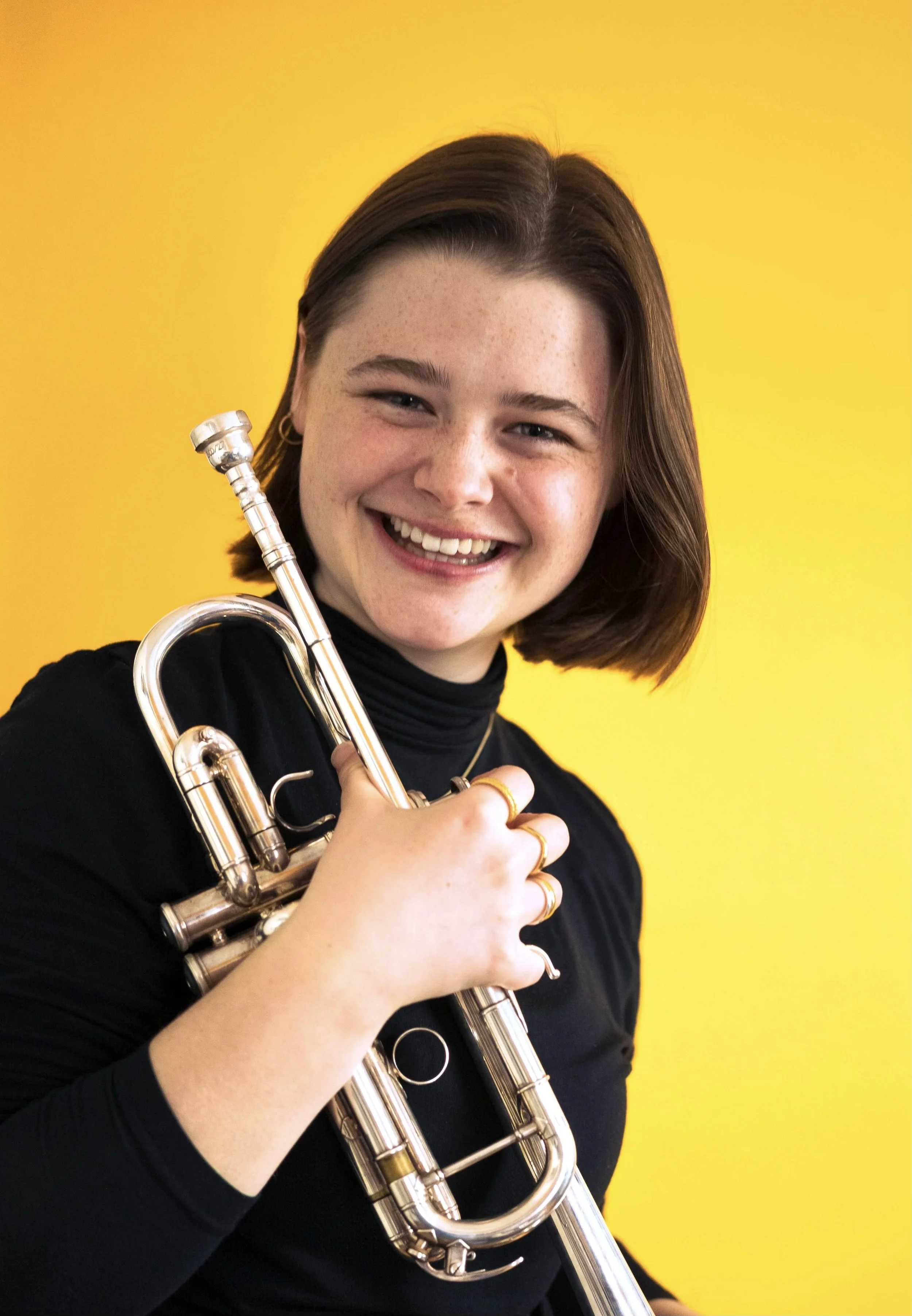 A young woman with short brown hair smiling while holding a silver trumpet against a yellow background.
