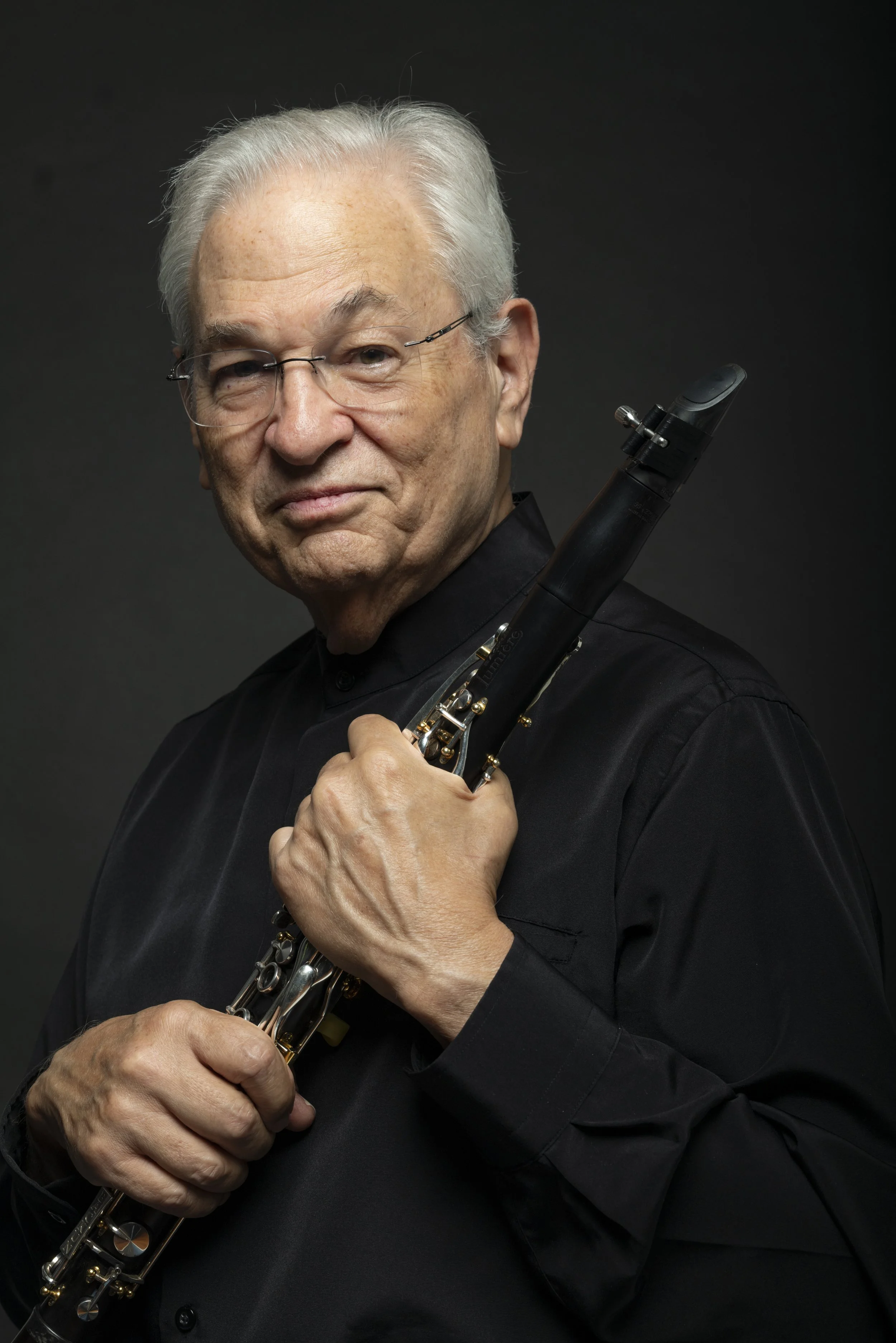 An elderly man with gray hair and glasses holding a clarinet against a dark background.
