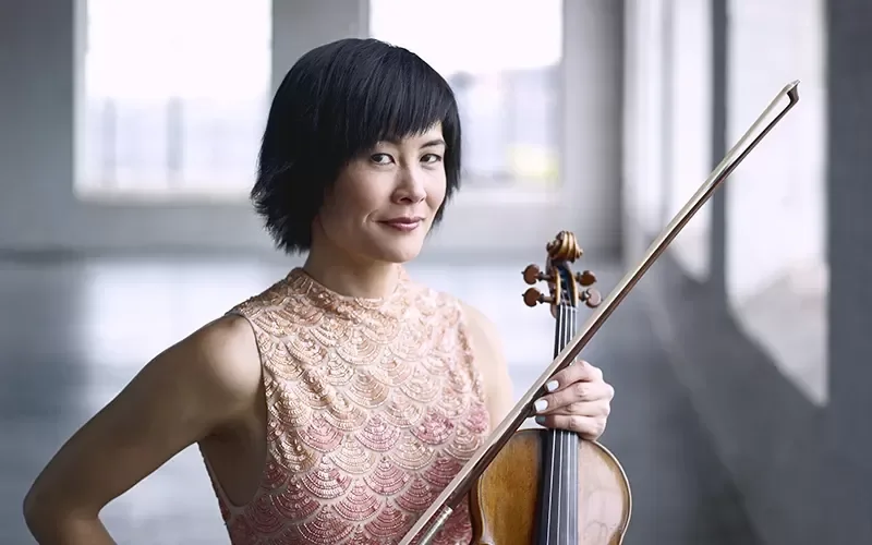 Woman with short black hair holding a violin and bow in a room with large windows.