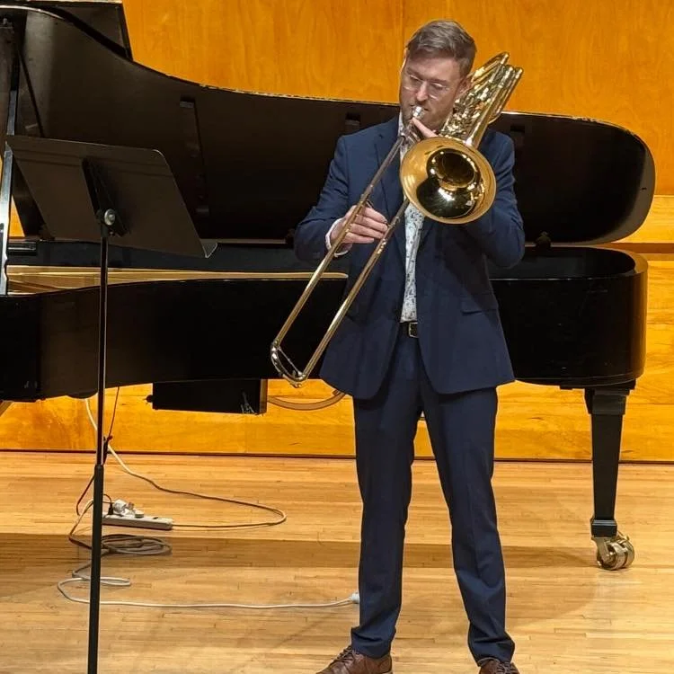 A man in a blue suit playing a trombone on stage with a black grand piano in the background.