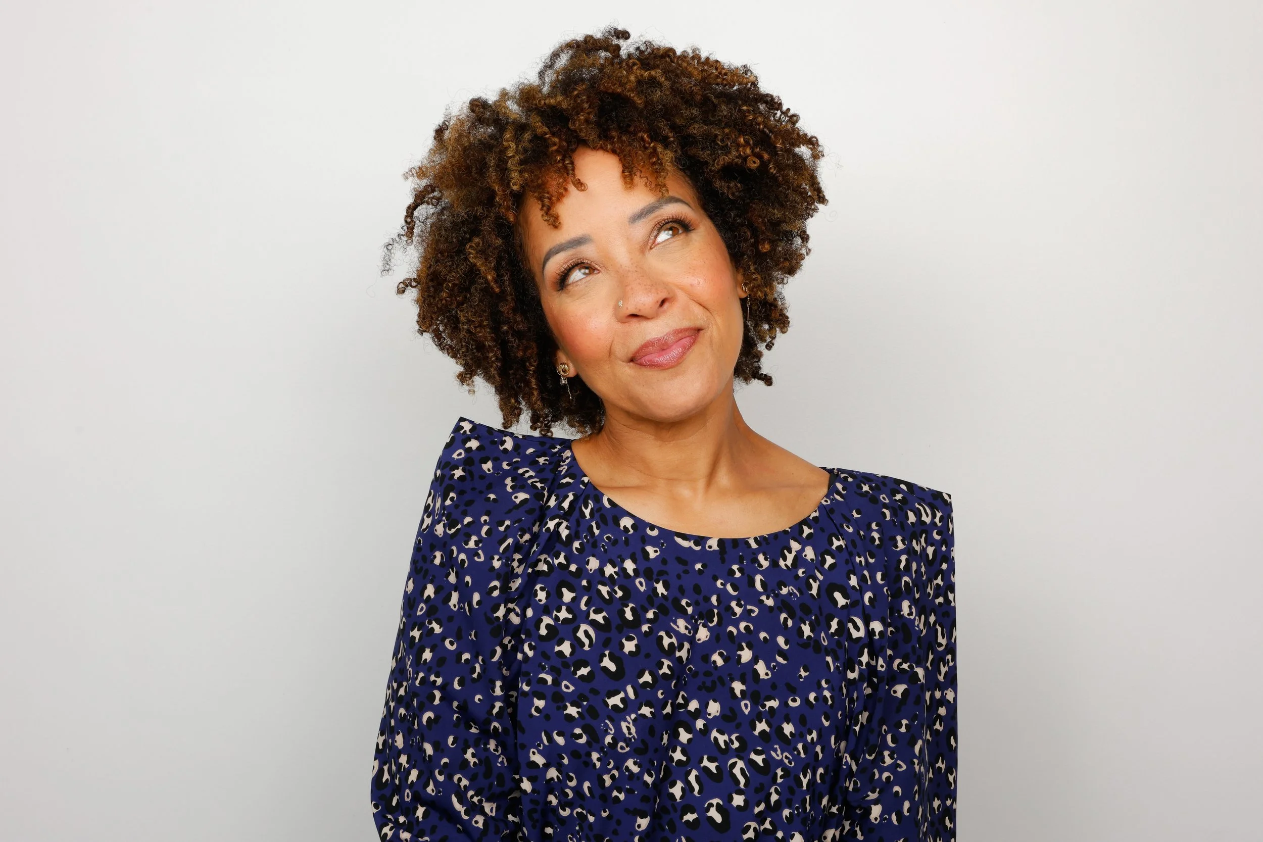 A woman with curly brown hair looking upward with a slight smile, wearing a blue animal print dress, standing against a plain white background.