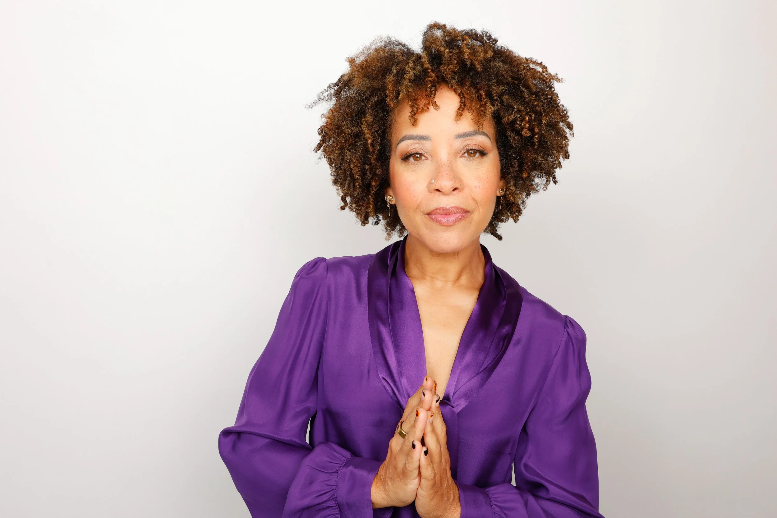 Woman with curly hair in a purple satin blouse standing against a plain background, hands pressed together in a prayer gesture, looking at the camera.