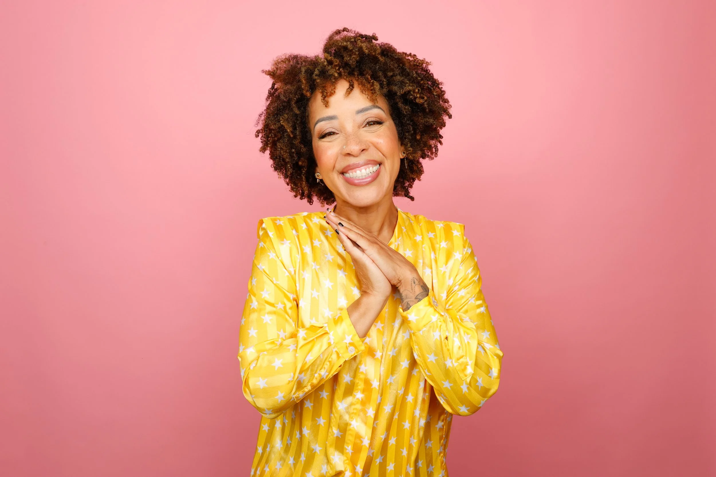 A woman with curly hair wearing a yellow star-patterned dress, smiling with eyes closed, standing against a pink background.