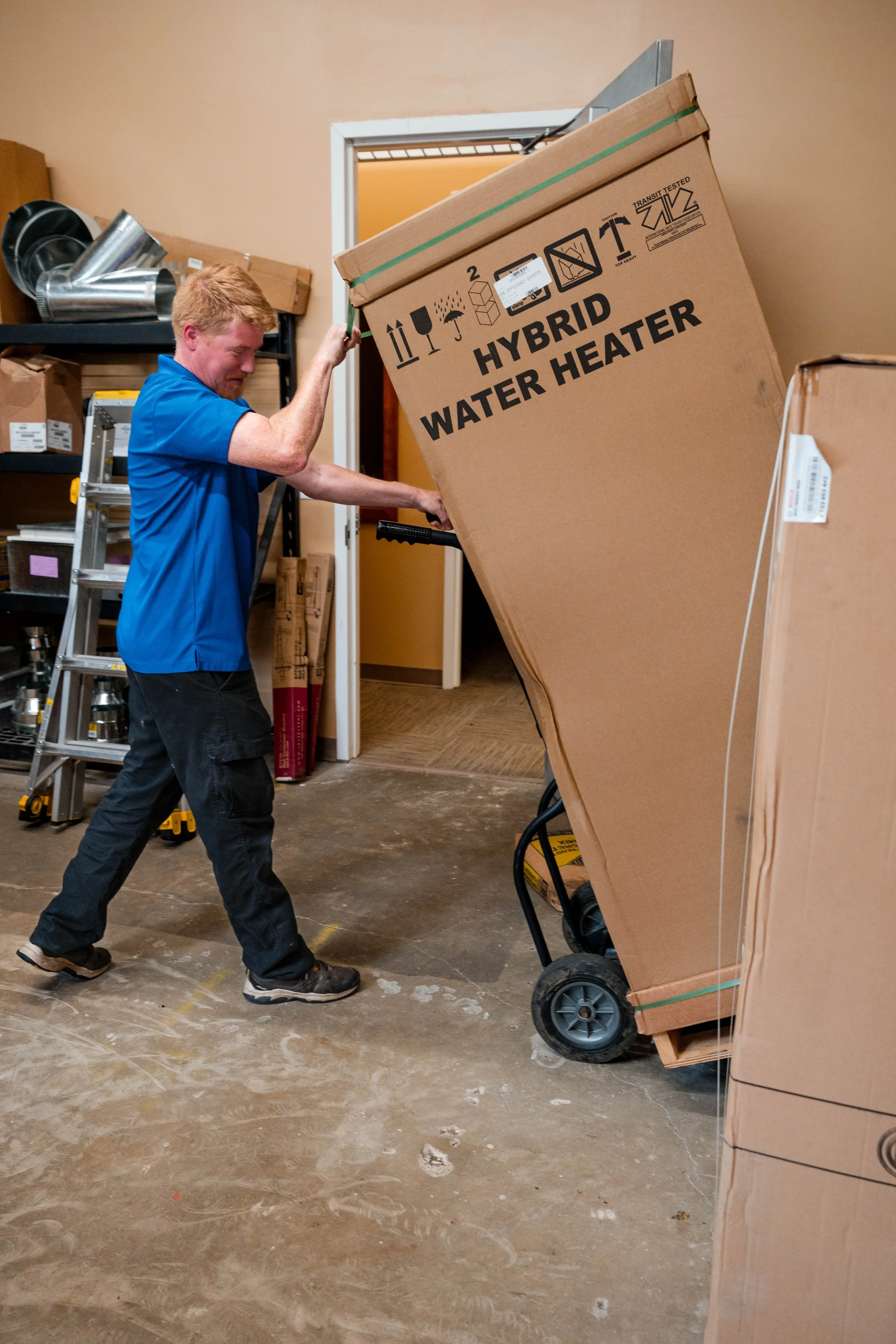 A man wearing a blue shirt and black pants is pushing a large cardboard box on a hand truck. The box is labeled 'HYBRID WATER HEATER' and appears to be in a storage room with shelves, tools, and other boxes.