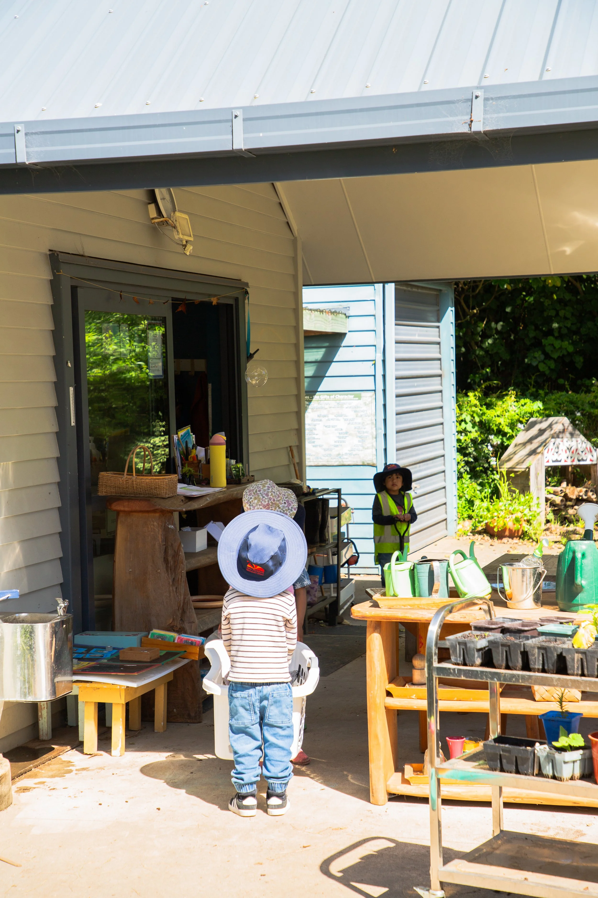 Children playing outdoors next to a house, with gardening tools and plants on tables, and a boy wearing a wide-brimmed hat and striped shirt facing a girl with a large sunhat, while another child stands in the background.