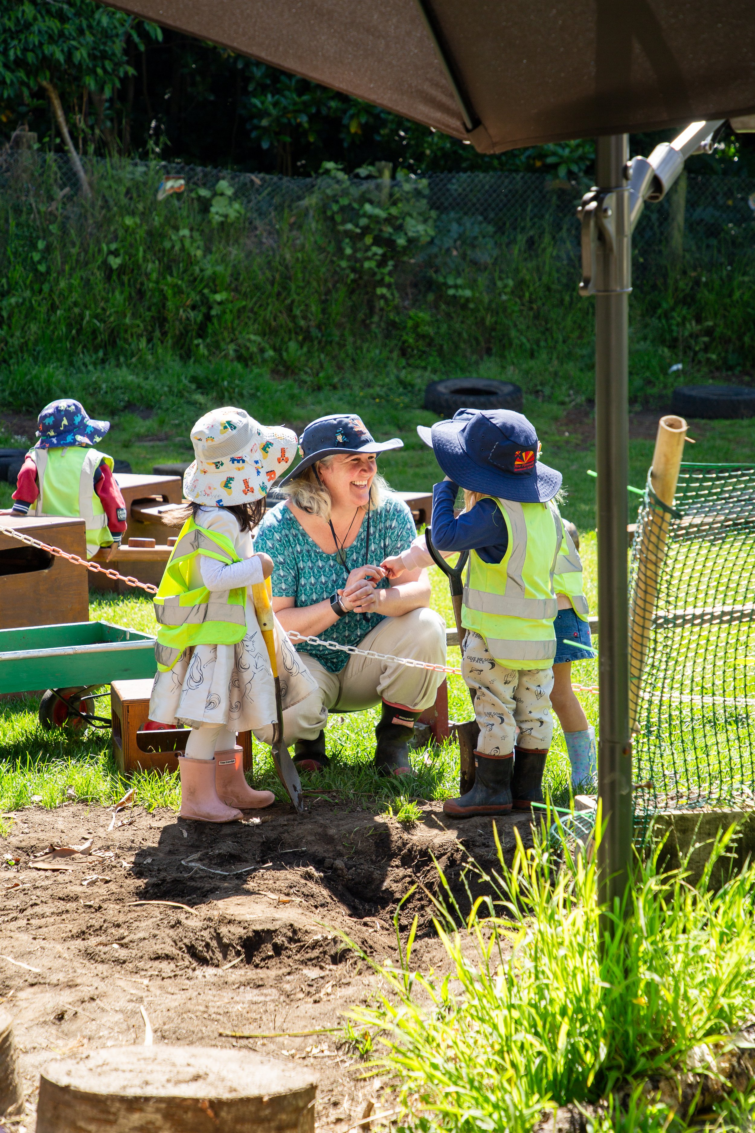 A woman and three children wearing sun hats and reflective vests are gathered around a small hole in the ground outdoors, with the woman smiling.