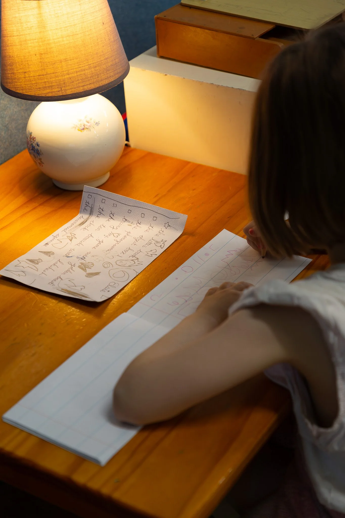 A person writing on lined paper at a wooden desk with a paper containing decorative writing nearby, a lamp with a floral design, and a white and wooden box in the background.