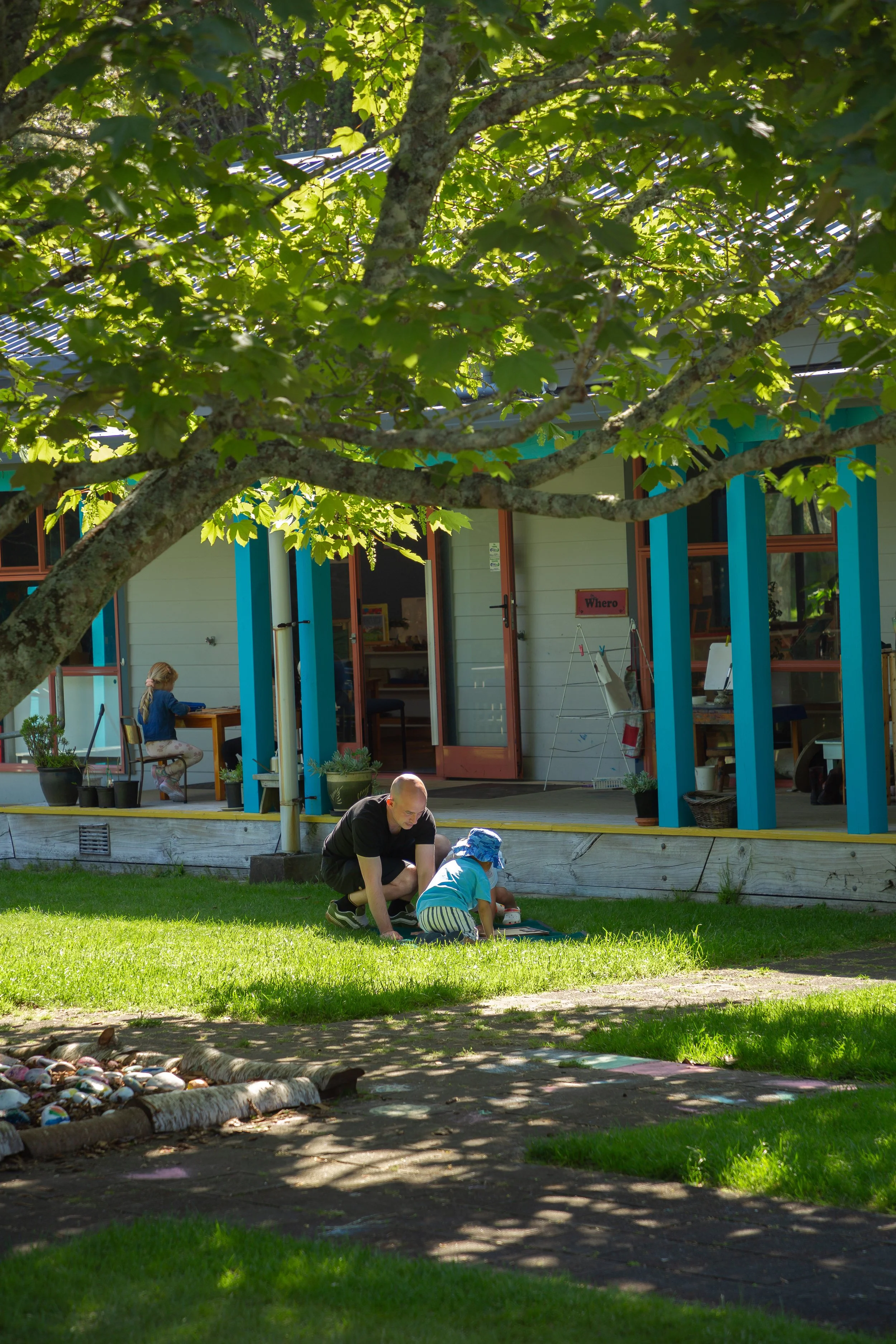A man and a young child playing on the grass outside a building with blue pillars, with another child sitting at a desk inside the building visible through an open door.