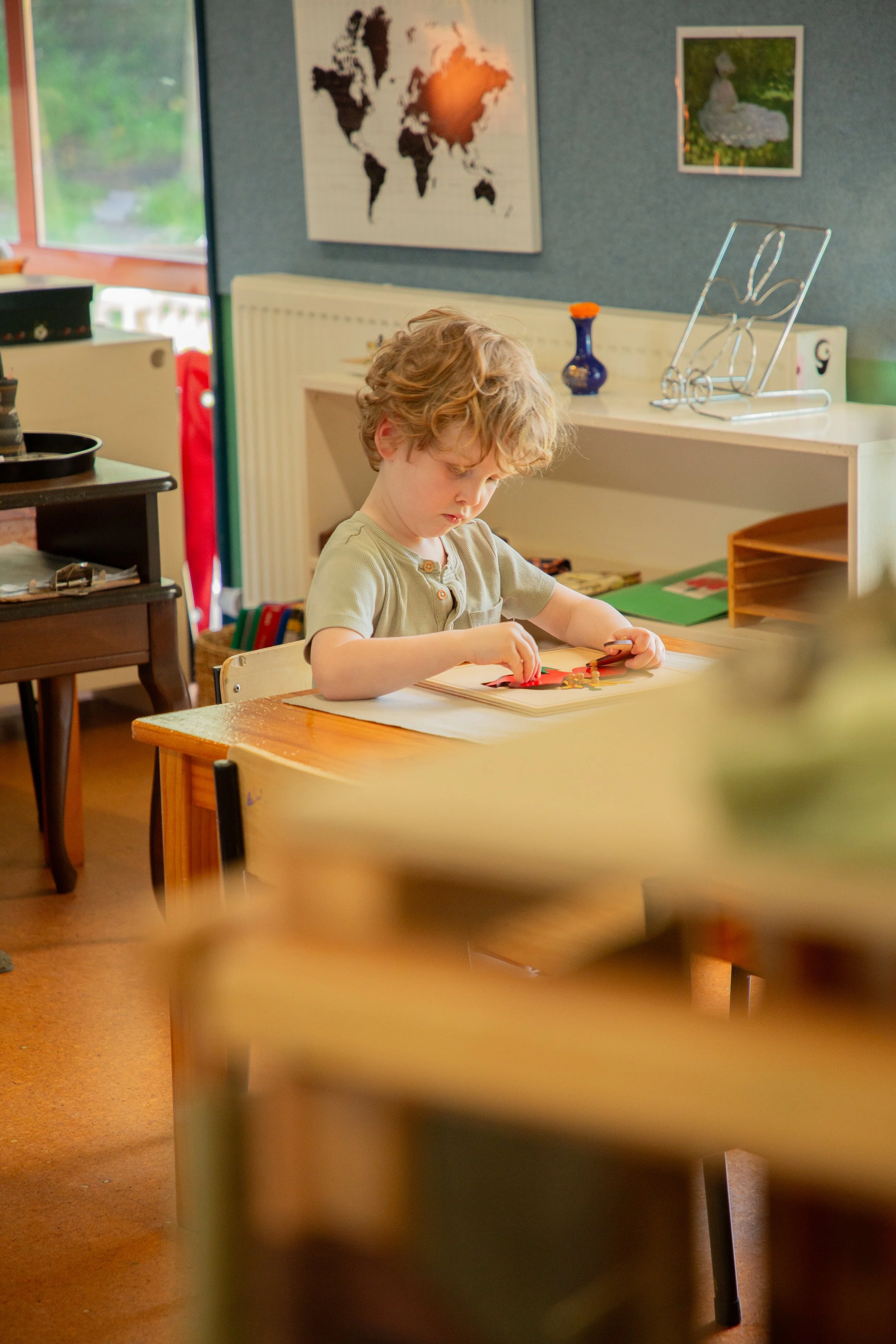 A young boy with curly blond hair sitting at a wooden table in a classroom, intently working on a puzzle.