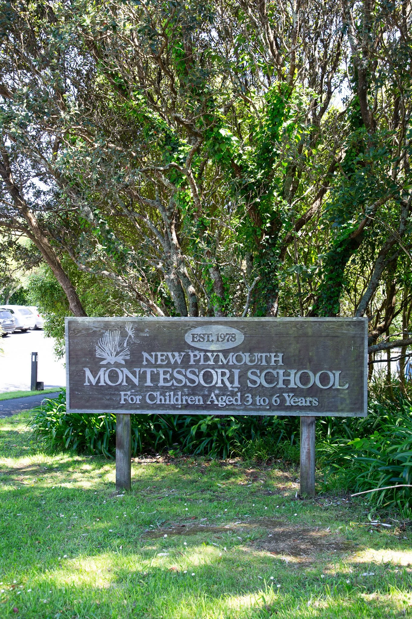 A wooden sign for New Plymouth Montessori School, established in 1978, located outdoors among trees and greenery. The sign indicates it is for children aged 3 to 6 years.