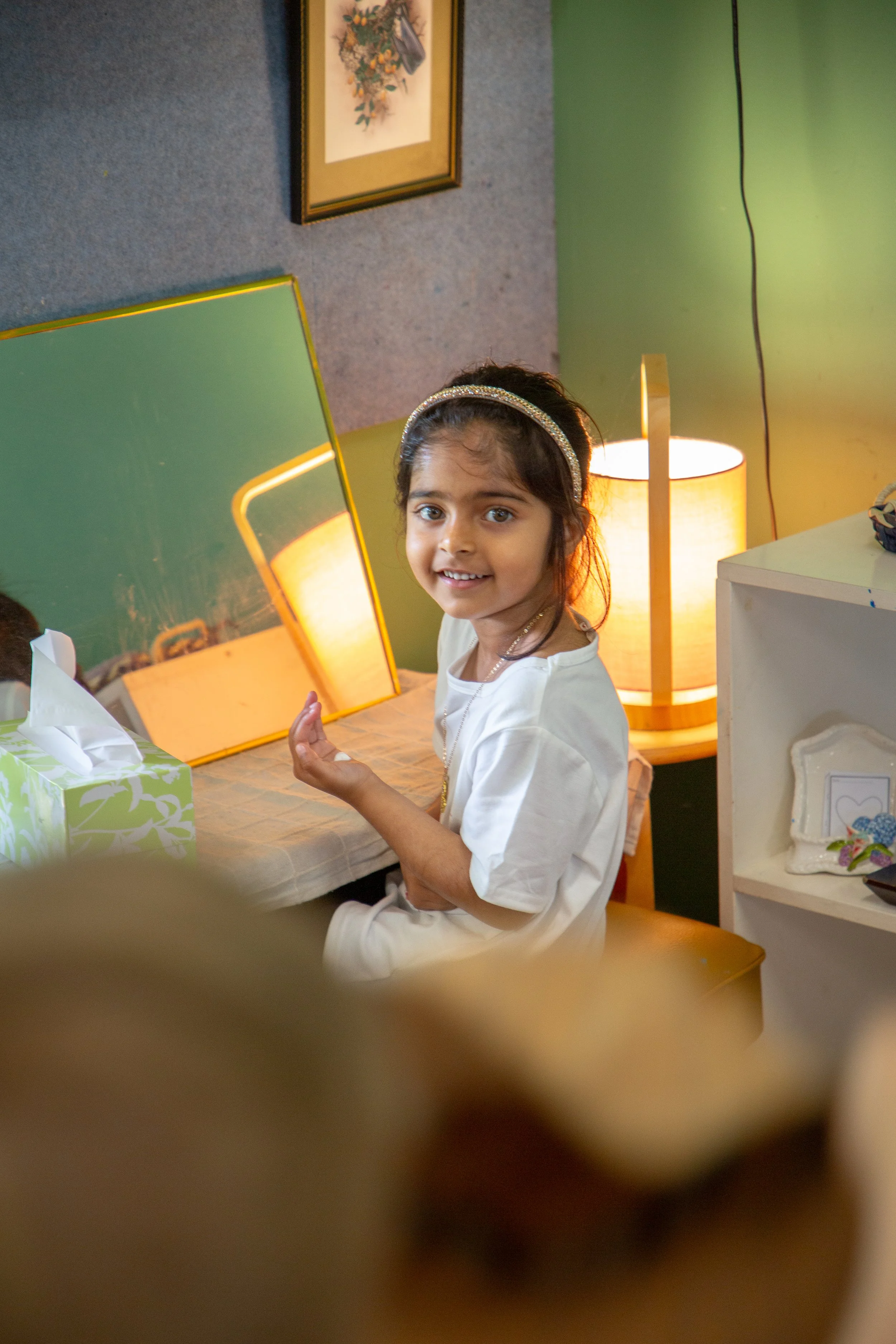 A young girl with dark hair wearing a white dress and headband, sitting at a table, smiling and looking at the camera. The background shows a lamp, framed artwork, and a green wall.