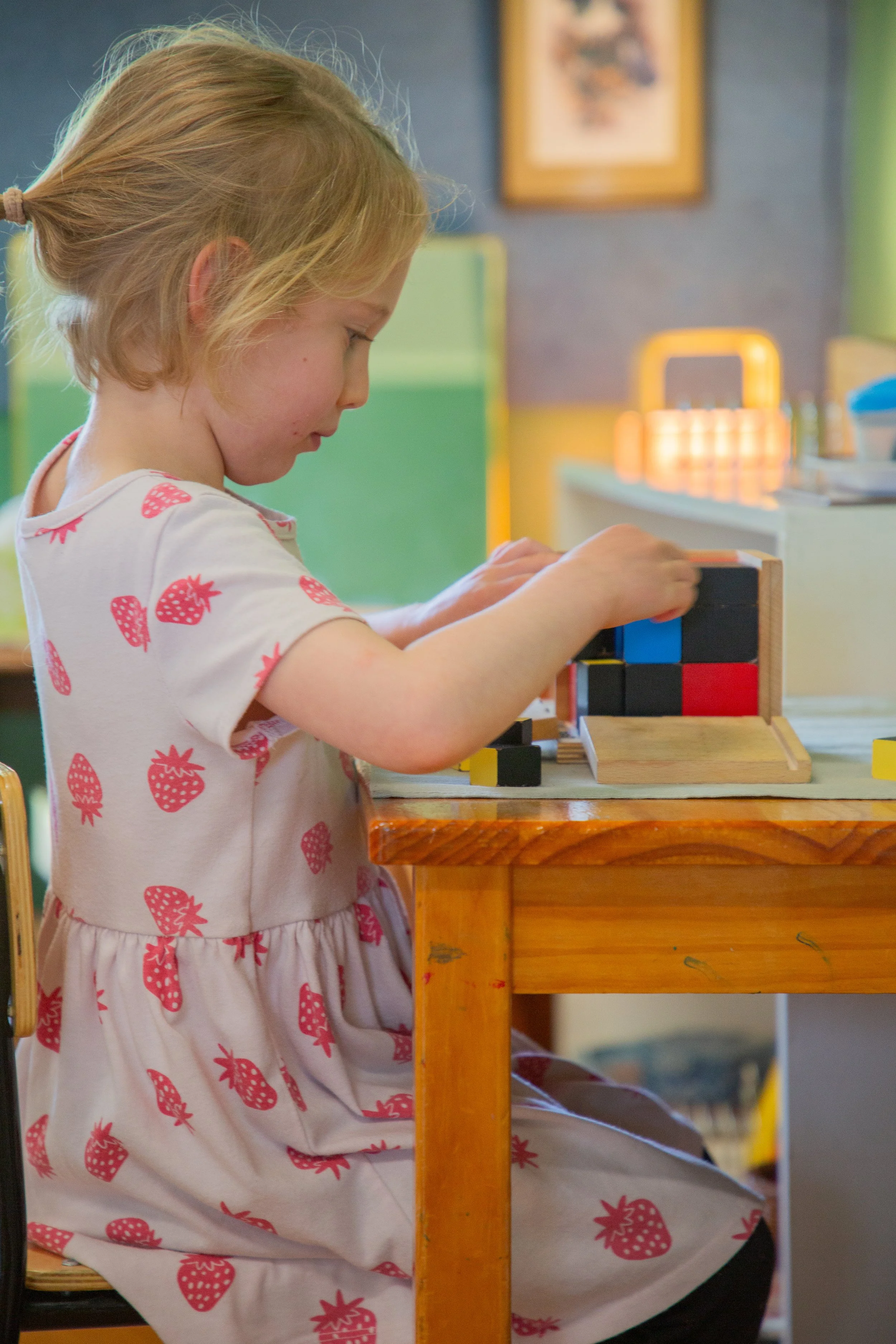 A young girl with blonde hair dressed in a white dress with pink strawberries, sitting at a wooden table and playing with stacking blocks.