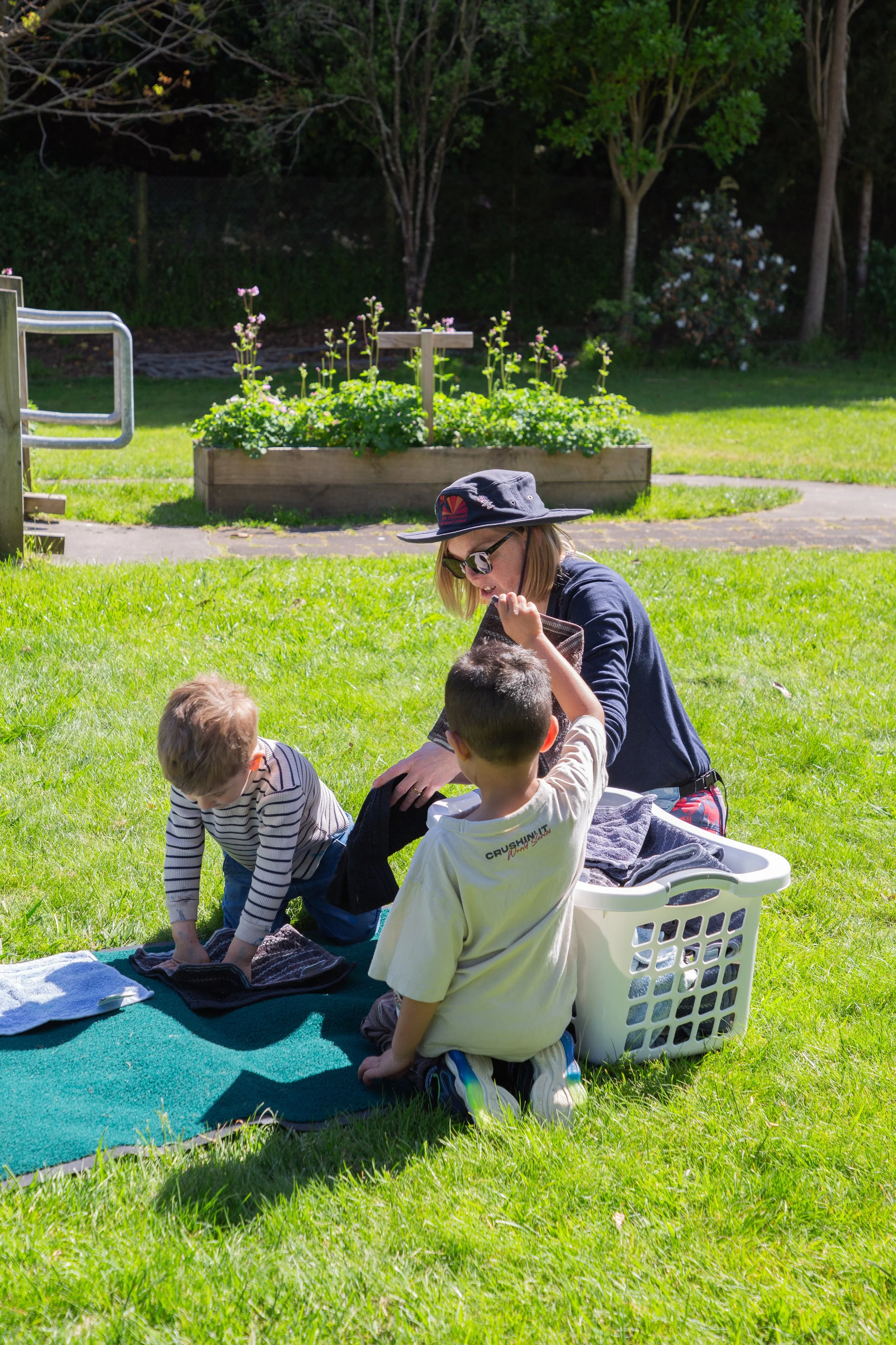 A woman and two young boys are outdoors on a grassy area with trees and a flower bed in the background. The woman is wearing sunglasses and a wide-brimmed hat, folding clothes or towels with the boys, who are kneeling on a green mat with laundry items.