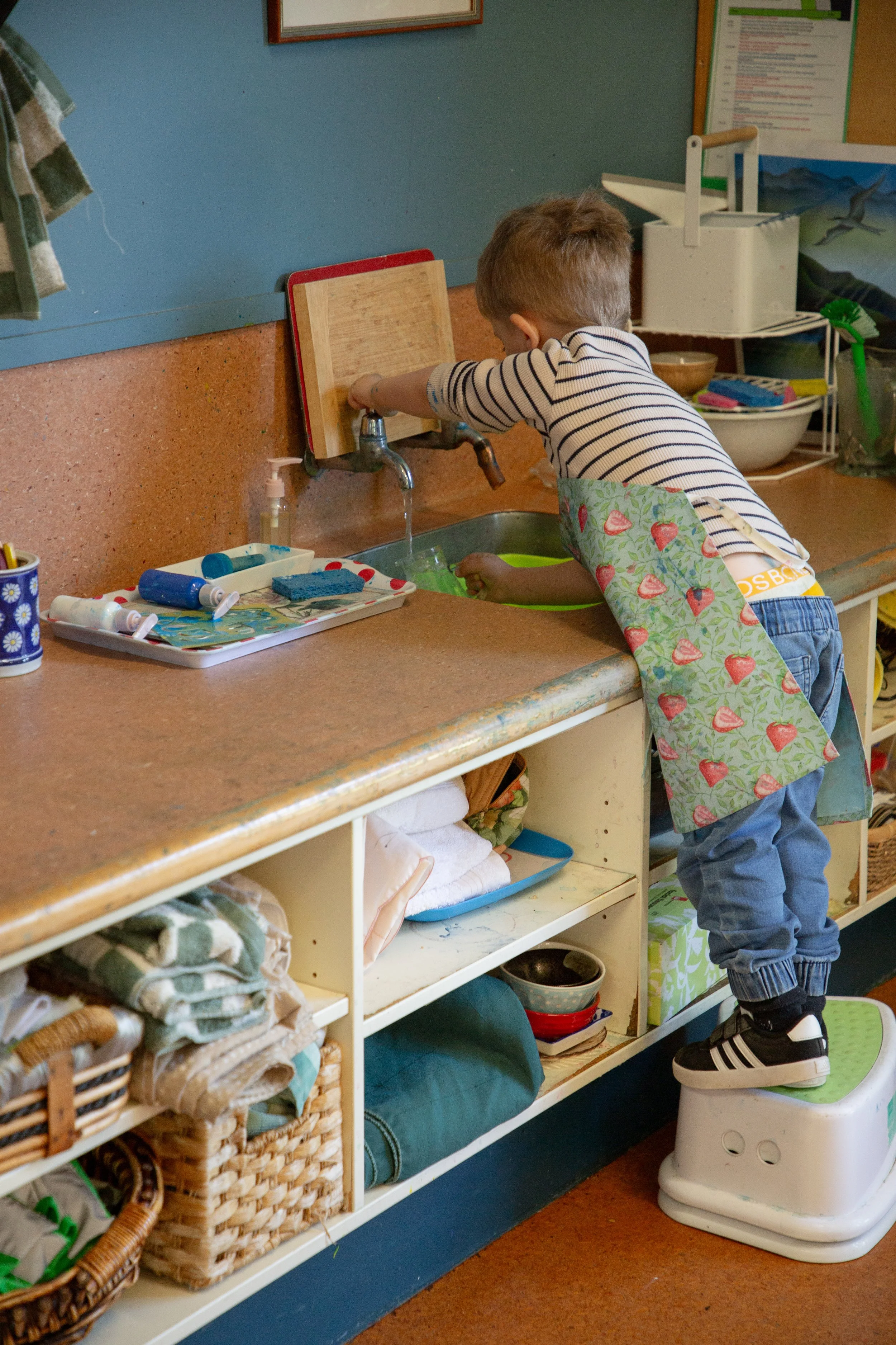 A young boy standing on a step stool at a kitchen sink, washing dishes, wearing an apron.