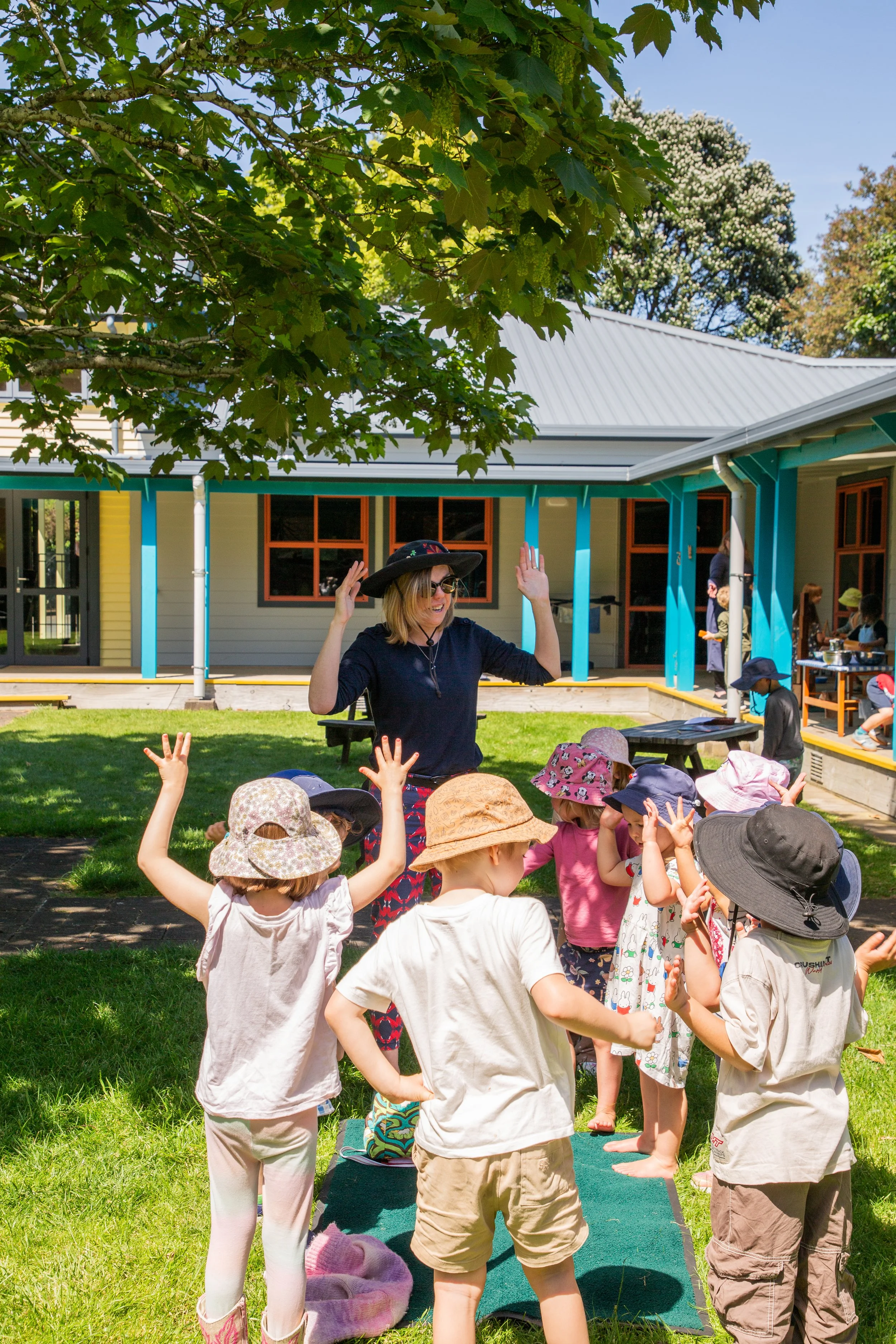 A woman wearing sunglasses, a black hat, and dark clothes leading a group of young children in an outdoor activity under a large leafy tree. Some children are wearing hats and casual summer clothes, standing on a green mat on the grass. The background shows a building with a gray roof, blue-turquoise support columns, and windows, with other children and adults visible on a deck.