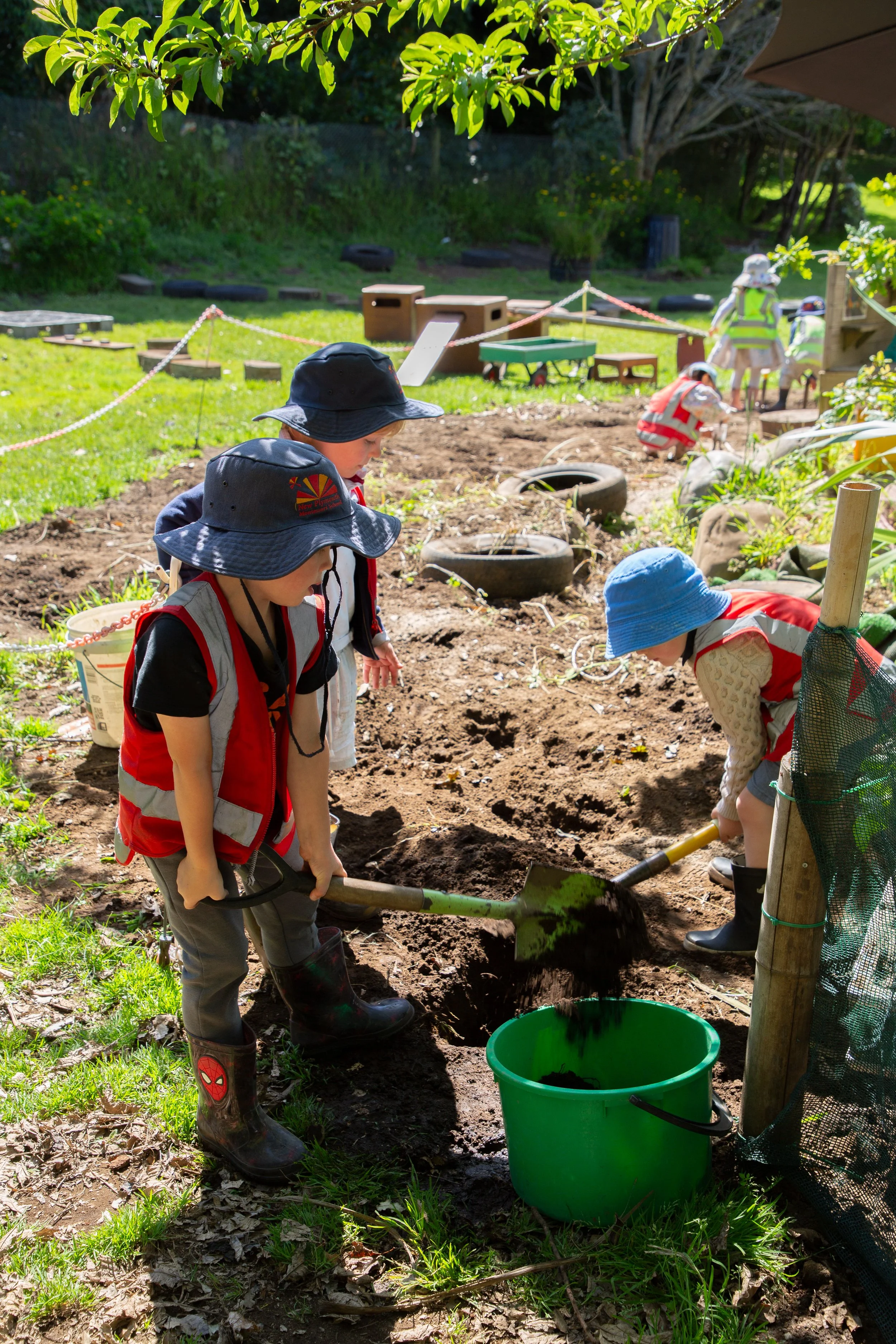 Children wearing hats and vests digging in dirt with shovels in a park or school yard, with other children in the background working on outdoor projects.