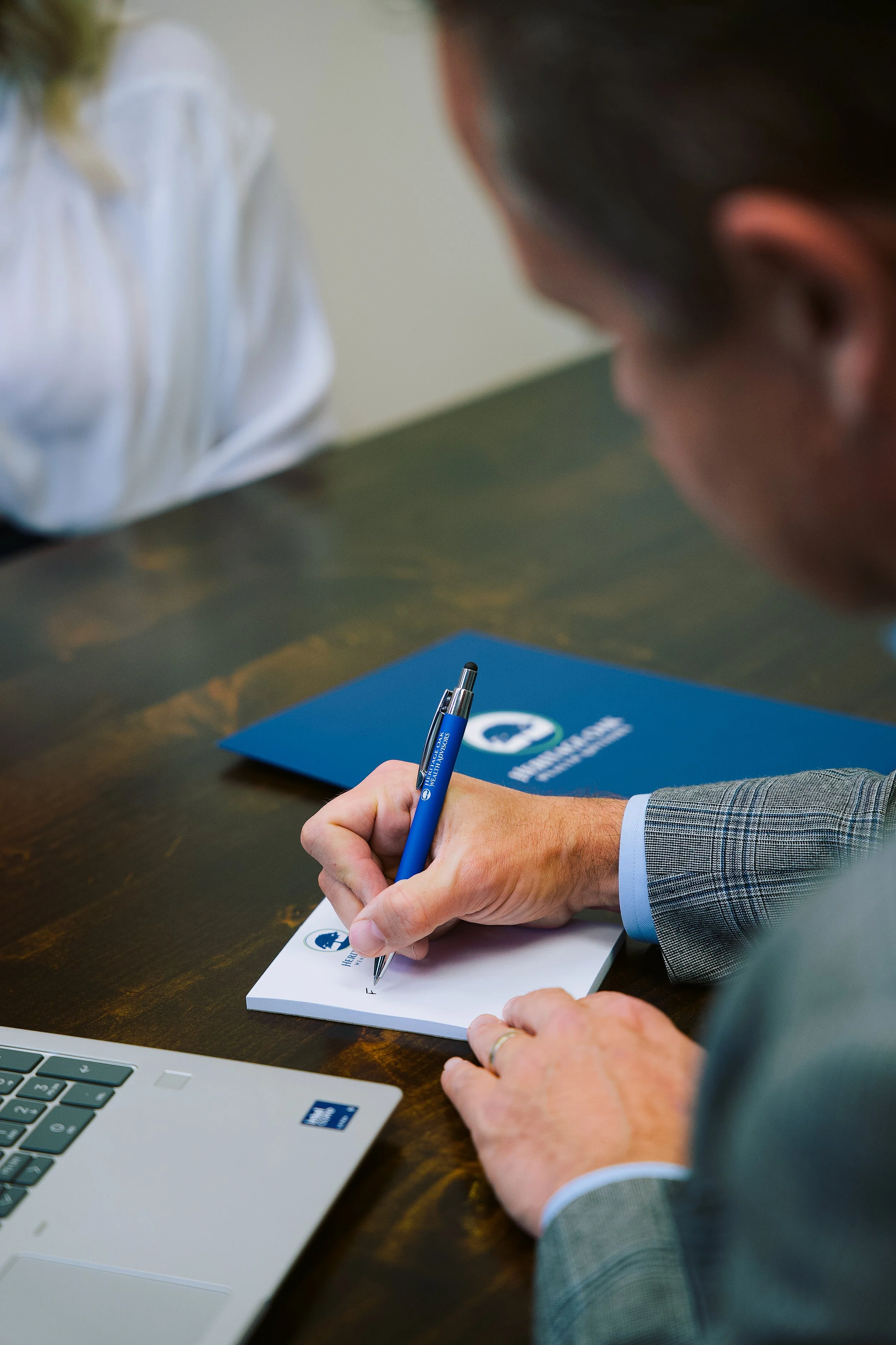 Person in a gray plaid suit signing a notepad with a blue pen at a conference table. A laptop and a blue folder with a logo are on the table.
