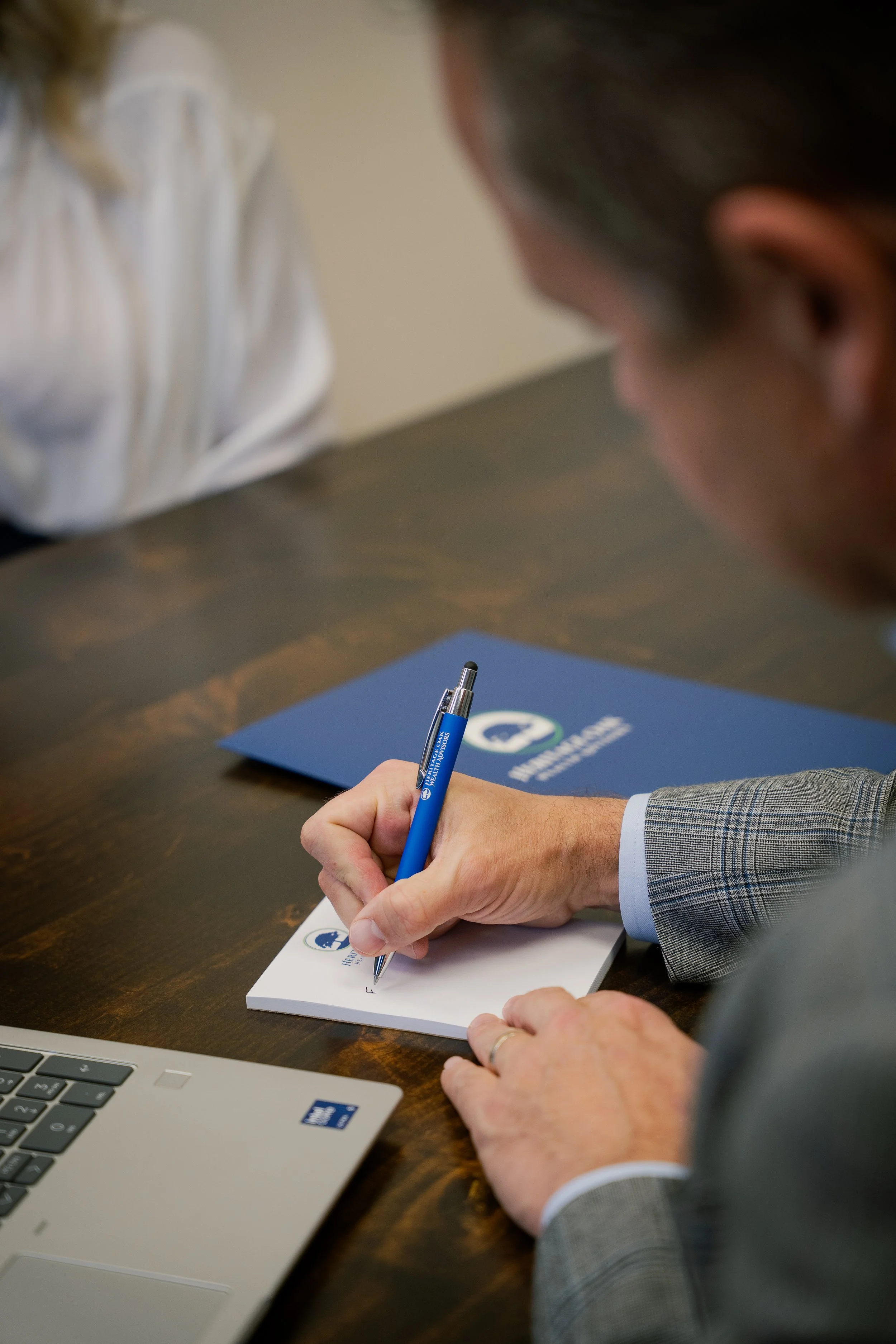 A person in a gray blazer signing a notepad with a blue pen at a wooden table. A laptop and a blue folder are also on the table.