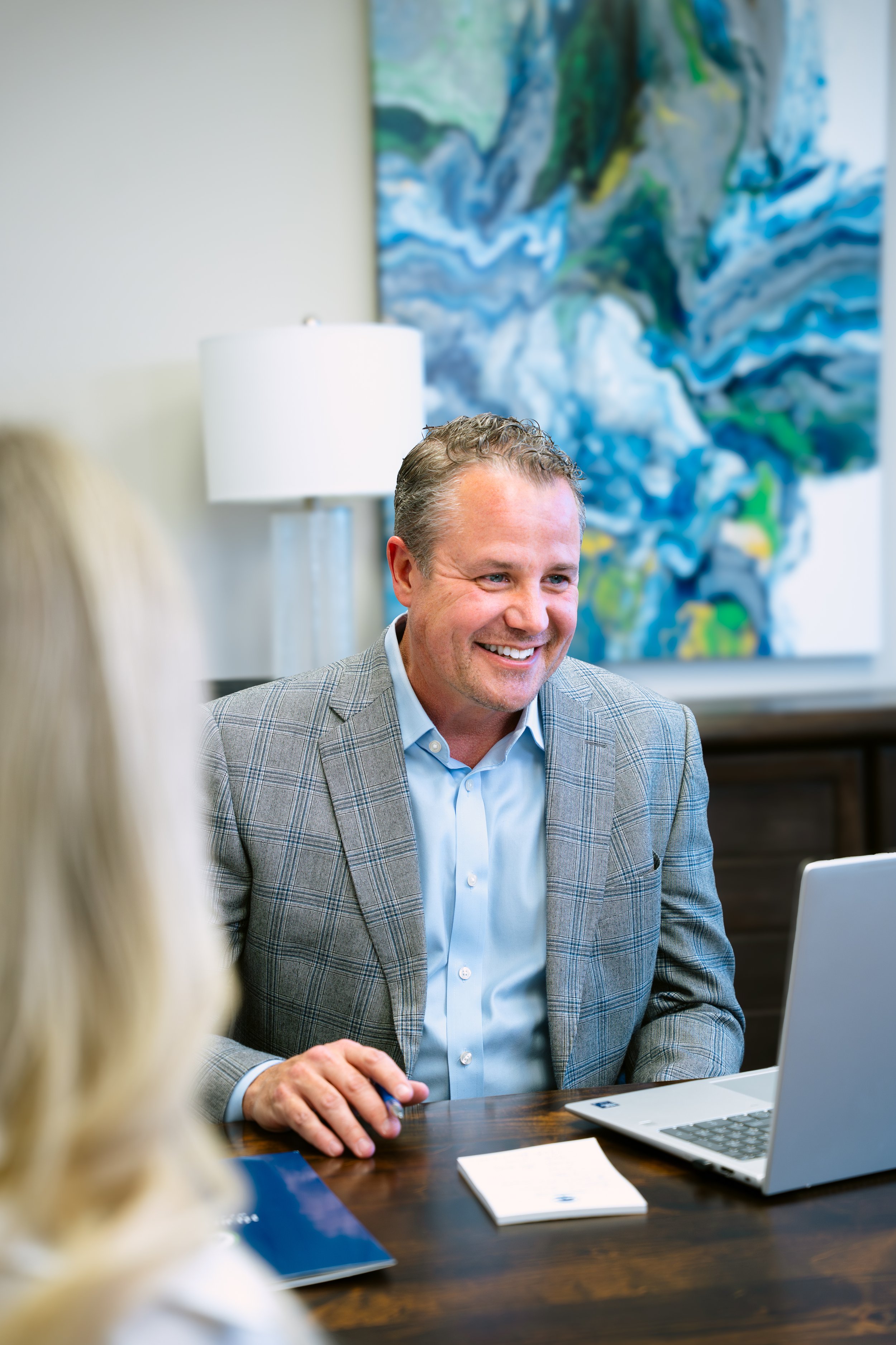 A man in a gray checked blazer smiles during a video conference in an office, with a woman with blonde hair visible in the foreground, a white lamp, and an abstract painting in the background.