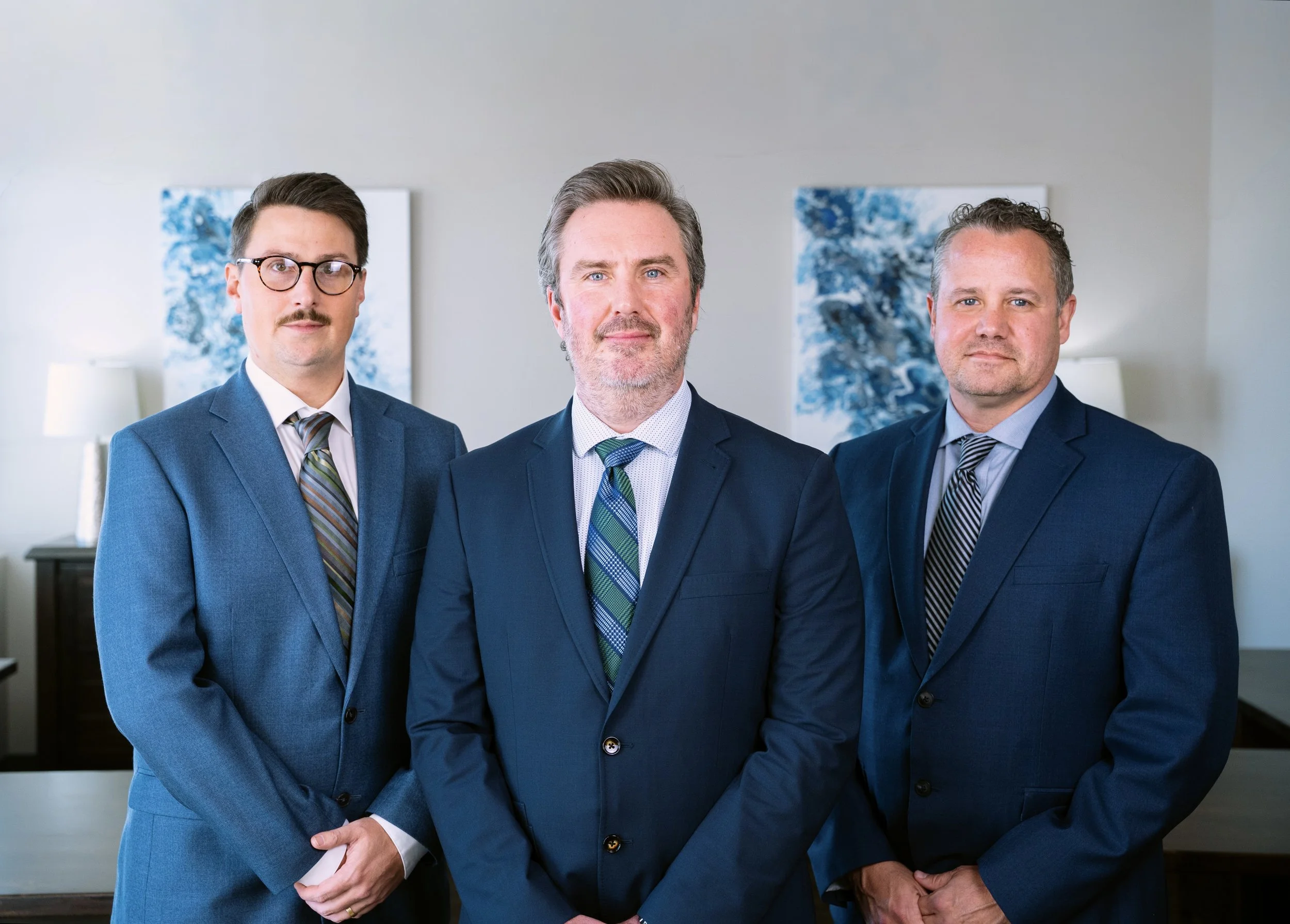 Three men in business suits standing in an office with abstract artwork and lamps in the background.