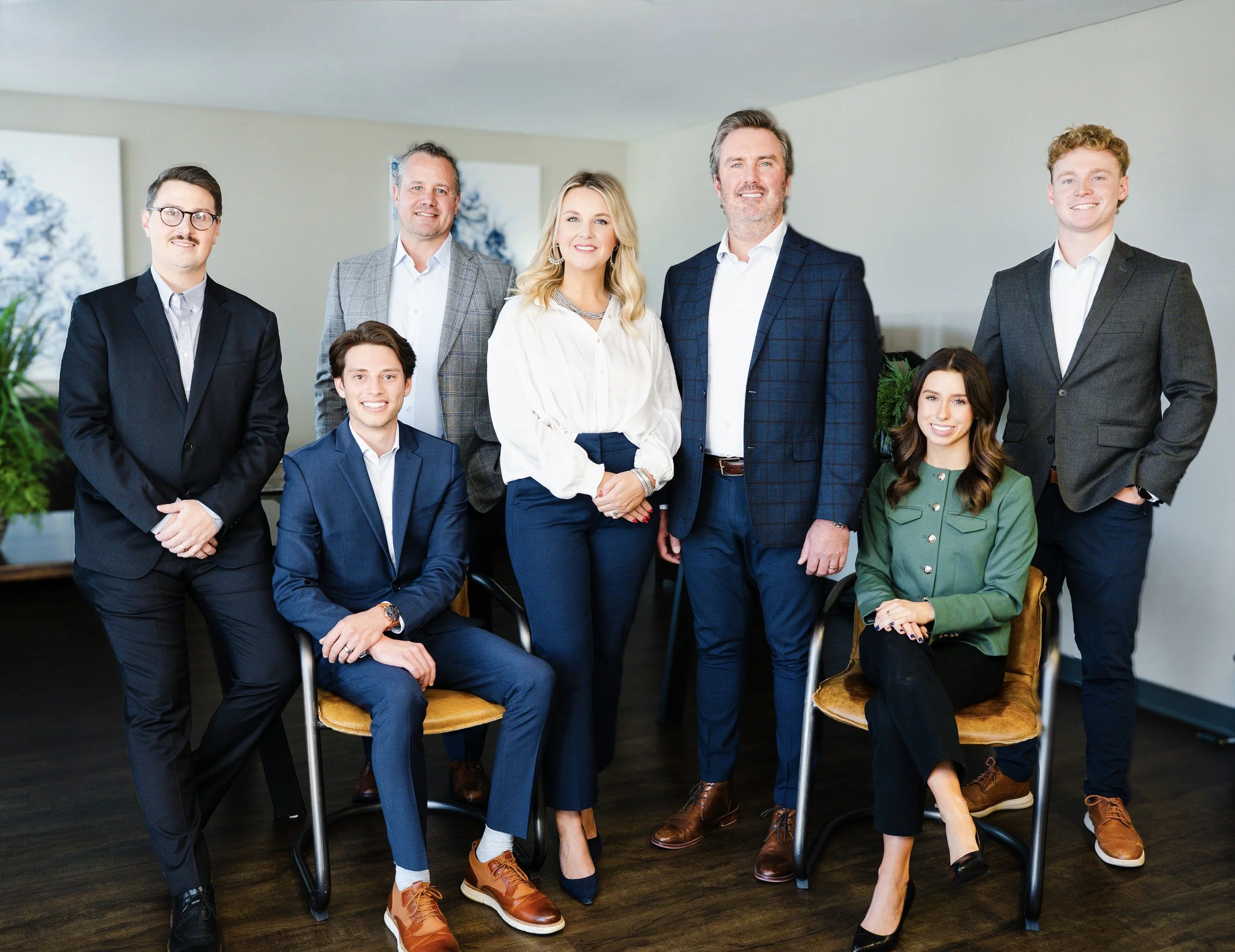 A professional group photo of eight people, five men and three women, dressed in business attire, in an indoor office setting with artwork and plants in the background.