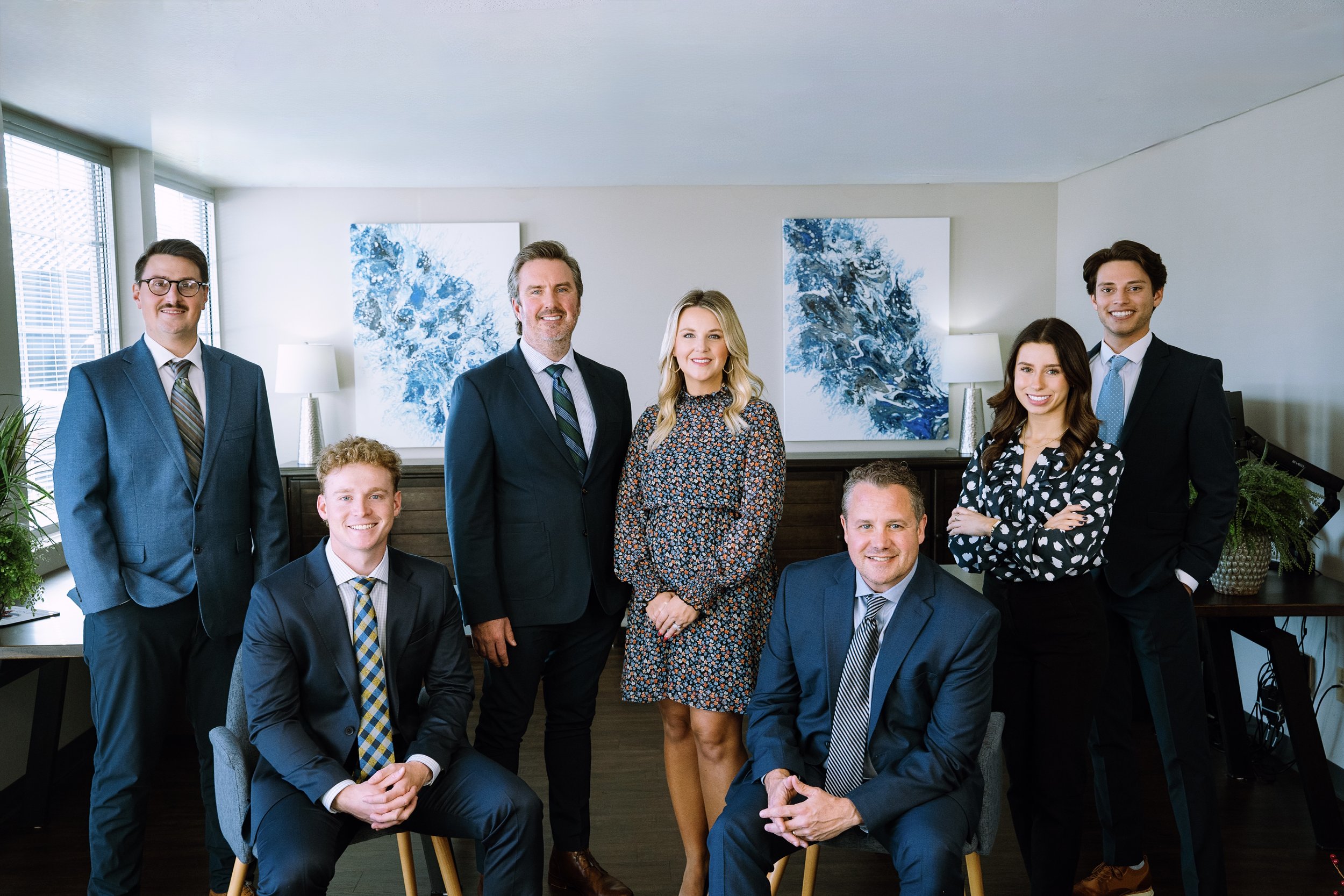 Group of eight professionally dressed people in an office, positioned in a semi-circle with some standing and some seated, smiling at the camera.