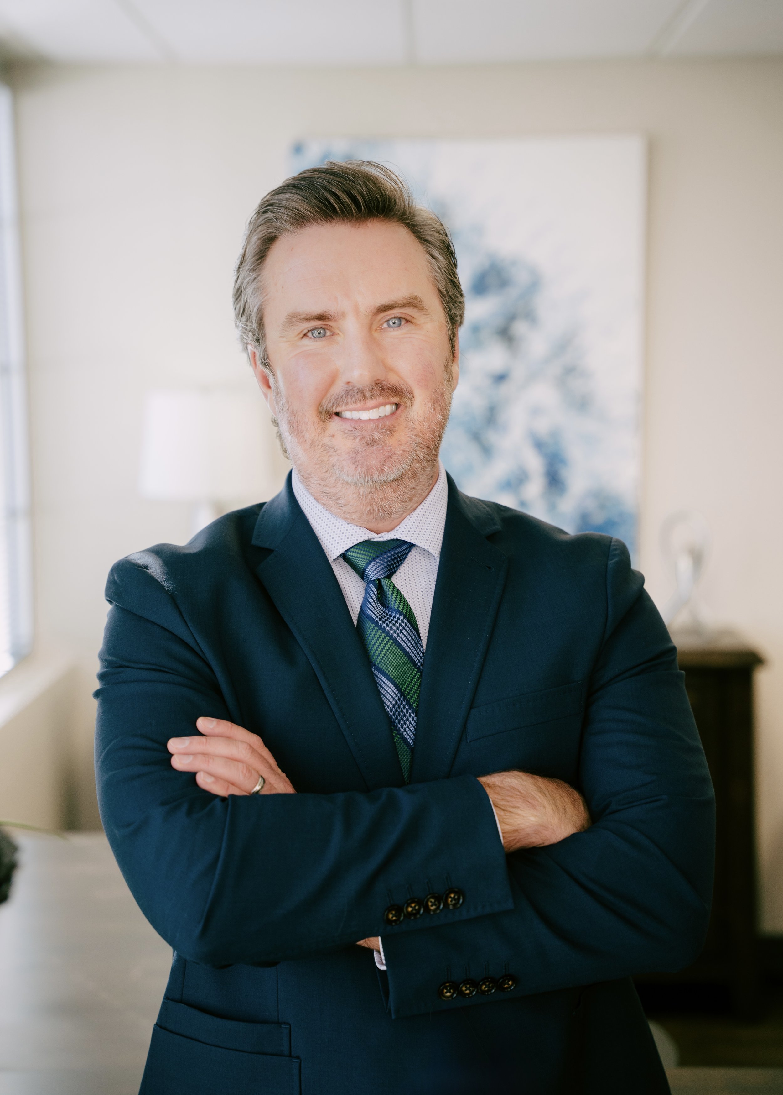 A smiling middle-aged man with grey hair and beard, wearing a navy suit, light blue shirt, and a blue-and-green striped tie, standing with arms crossed in a well-lit office or living room with a painting and a lamp in the background.