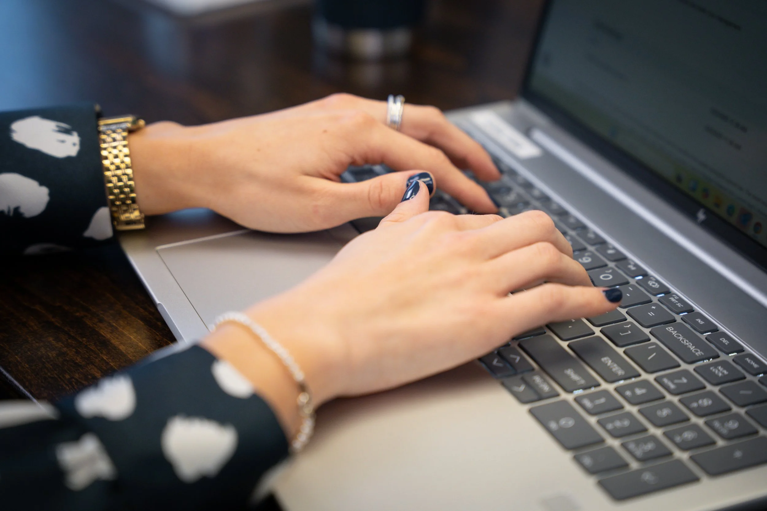 A person typing on a silver laptop with wearing a black watch with white patterns, a ring, and a bracelet, on a wooden table.
