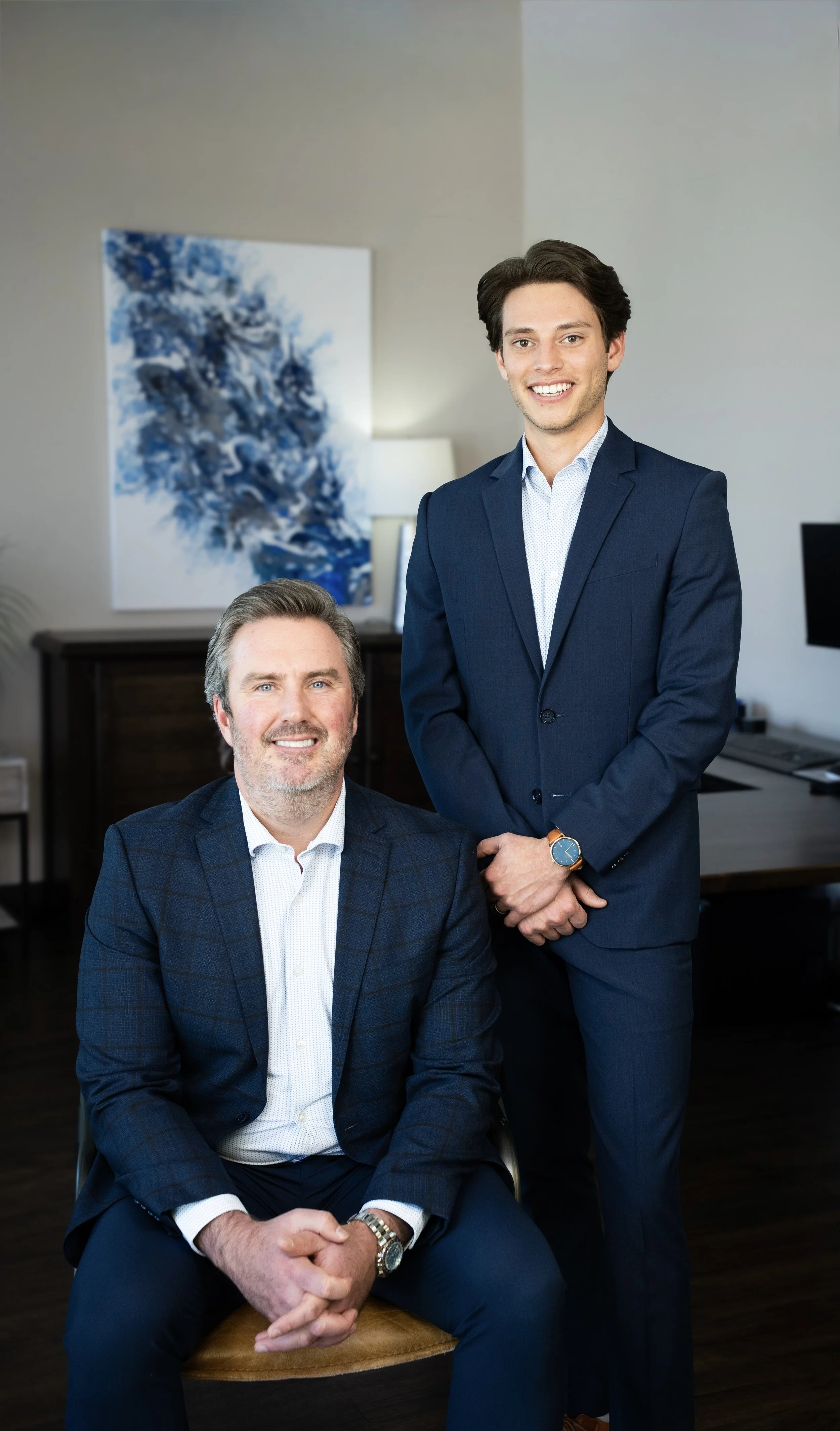 Two men in business suits smiling indoors, one seated and one standing, with a modern office background and blue abstract artwork on the wall.