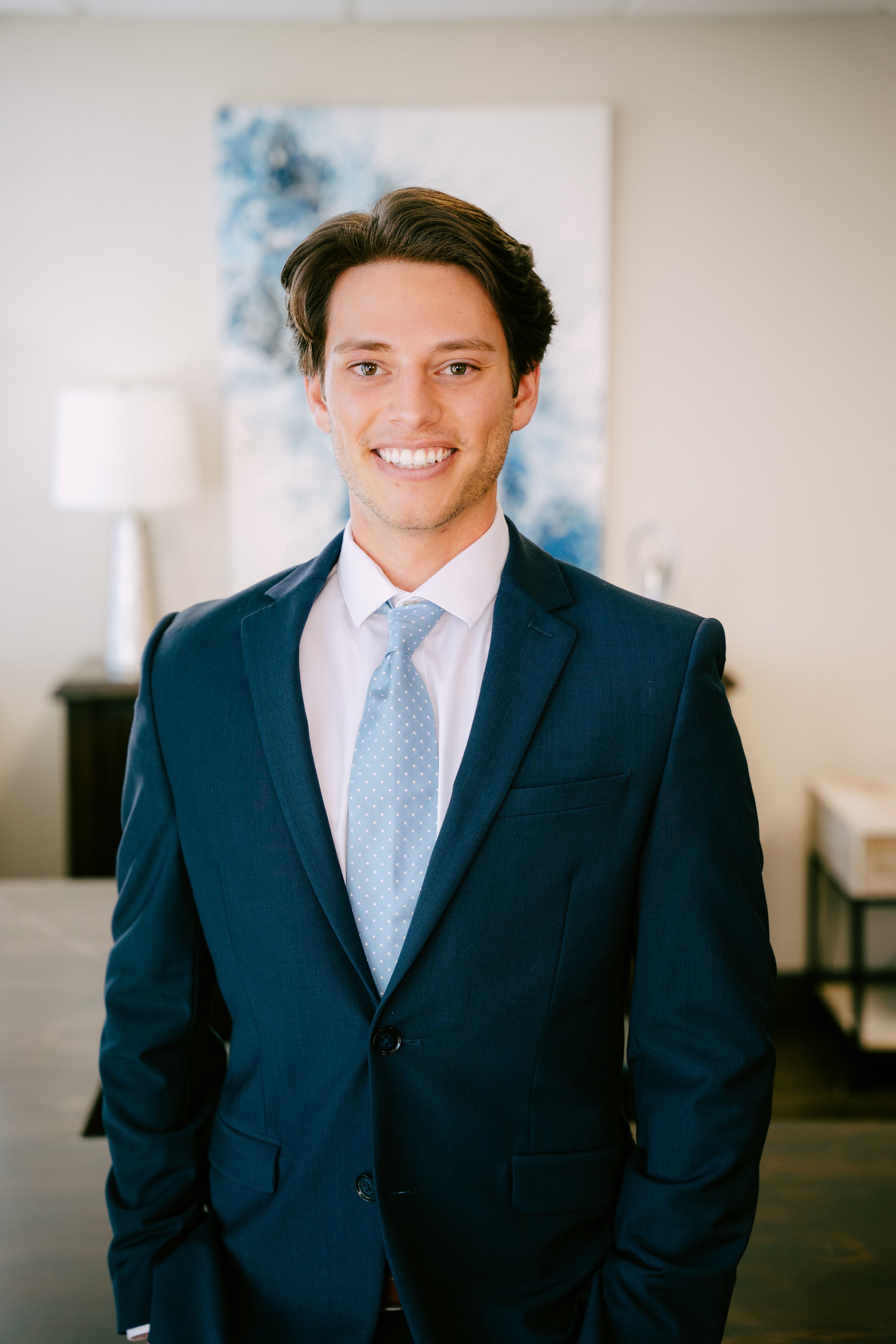A young man in a dark blue suit, white shirt, and light blue tie, smiling in a modern room with artwork on the wall and a lamp in the background.