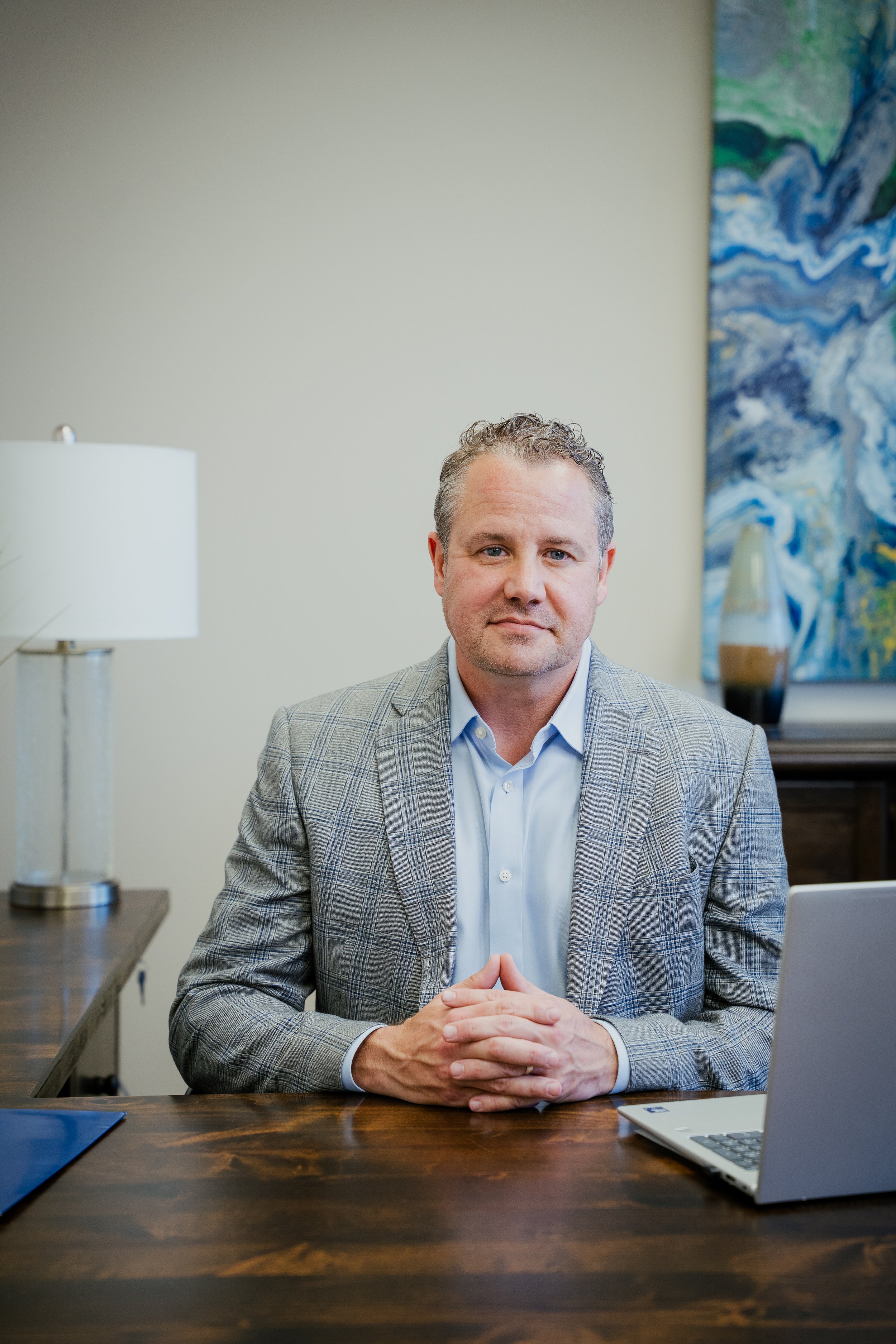 A man sitting at a wooden desk with a laptop and a lamp in a modern office with artwork and a vase in the background.