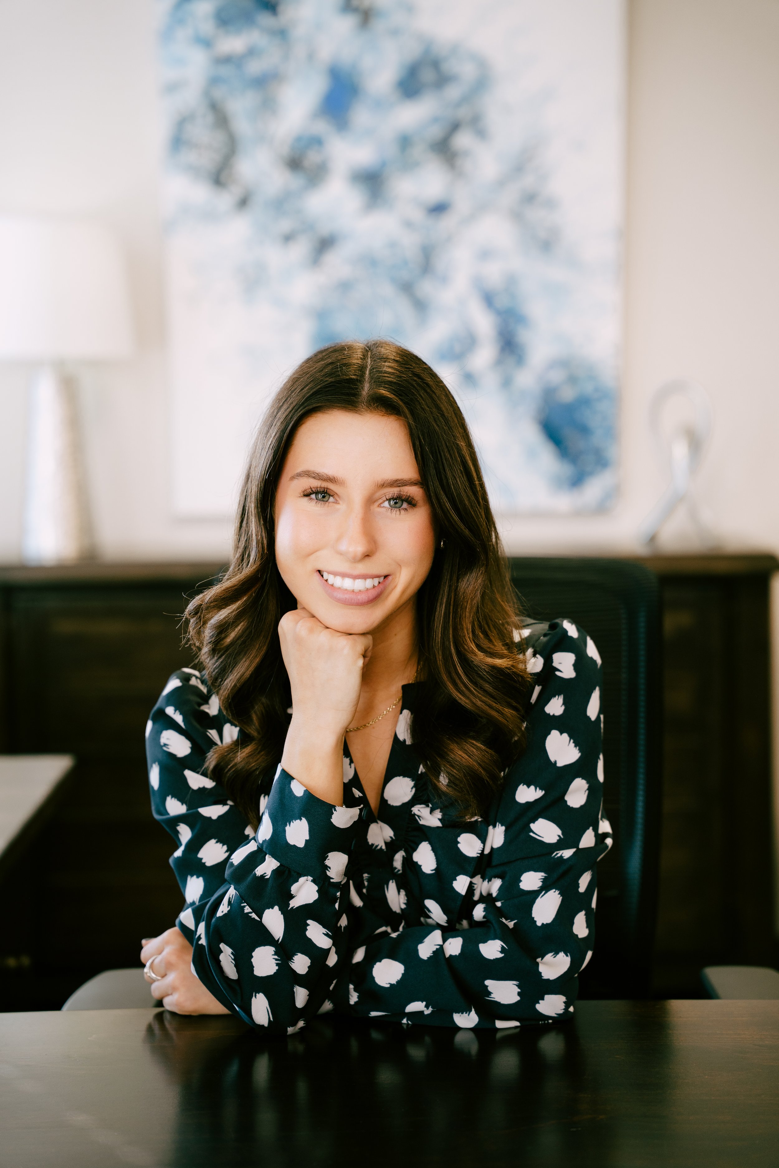 A woman with long dark hair and light skin, smiling, resting her chin on her hand, sitting at a desk in an office with abstract art in the background.