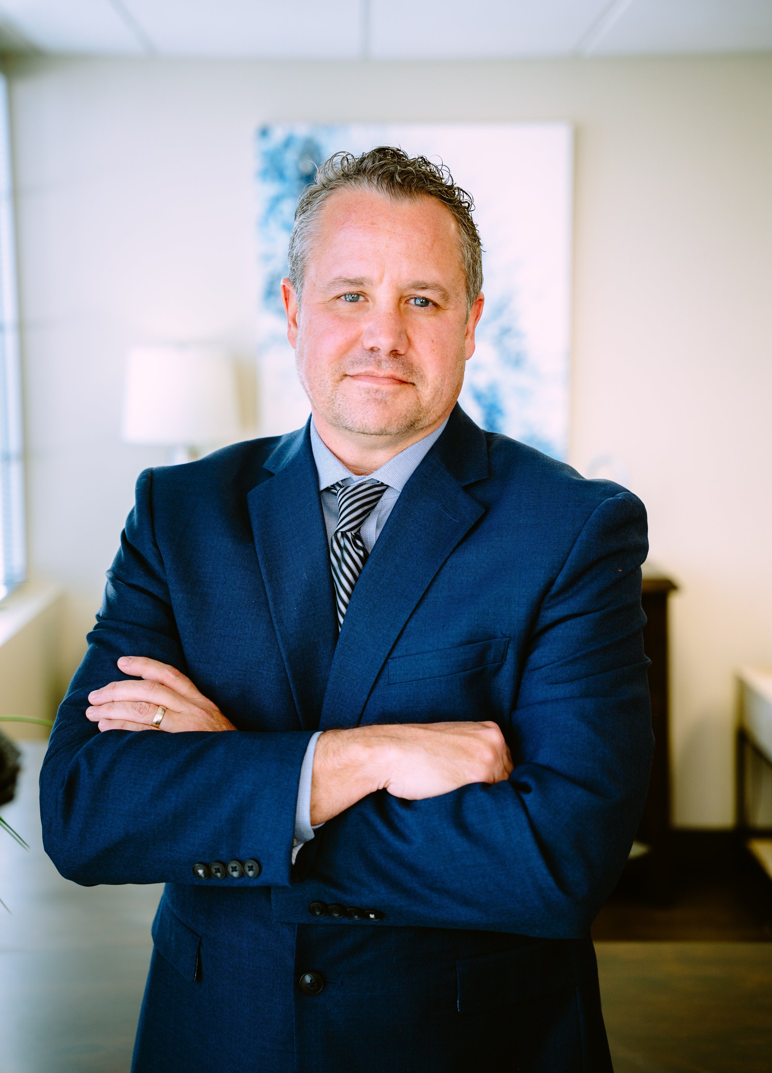 A man in a dark blue suit with arms crossed, standing in an office with light-colored walls, a window, and a framed abstract painting in the background.