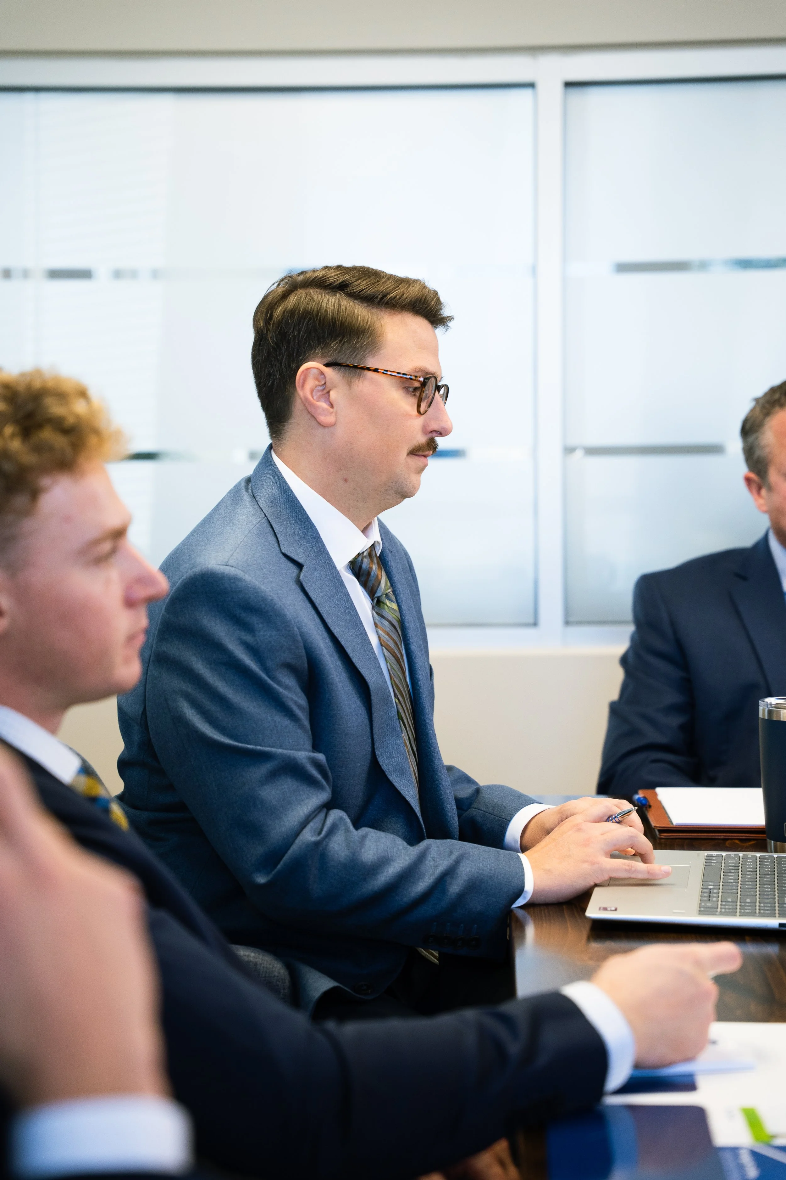 Three men in business suits sitting at a conference table during a meeting, with a laptop, notebooks, and a tumbler on the table.