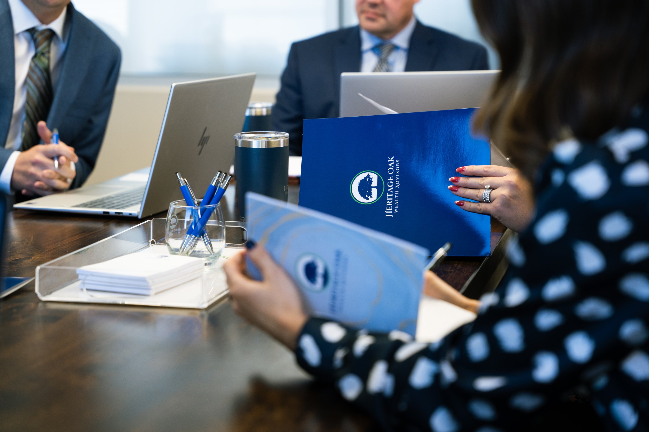 Business meeting with three people, laptops, pens, and brochures on a wooden table.