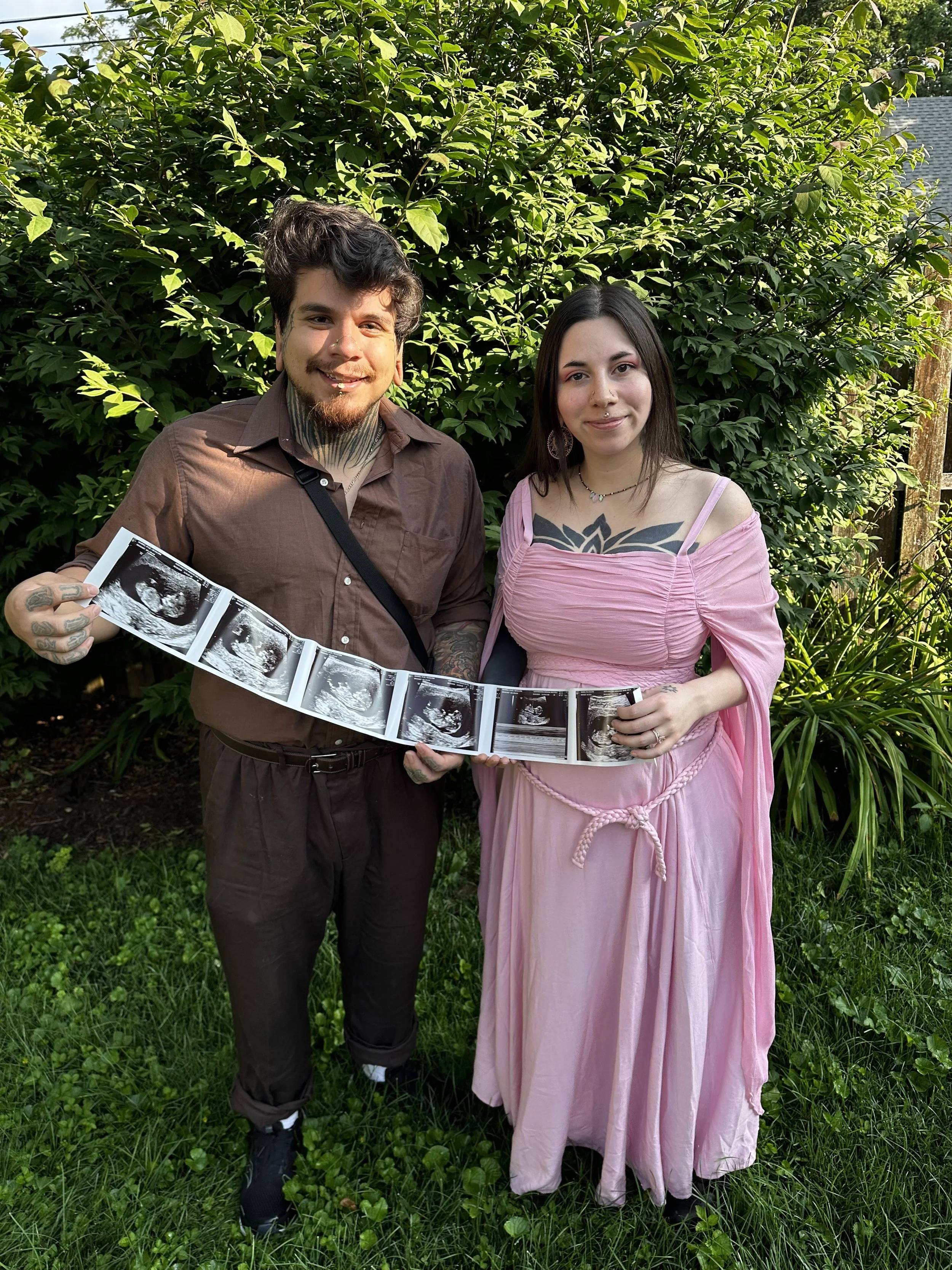 A young man and woman stand outdoors on grass, holding a string of ultrasound images, smiling at the camera. The man has tattoos and short hair, dressed in a brown shirt and pants. The woman has long dark hair, a septum piercing, wearing a pink dress with off-shoulder sleeves, and has visible tattoos on her chest and arms. The background features lush green bushes and trees.