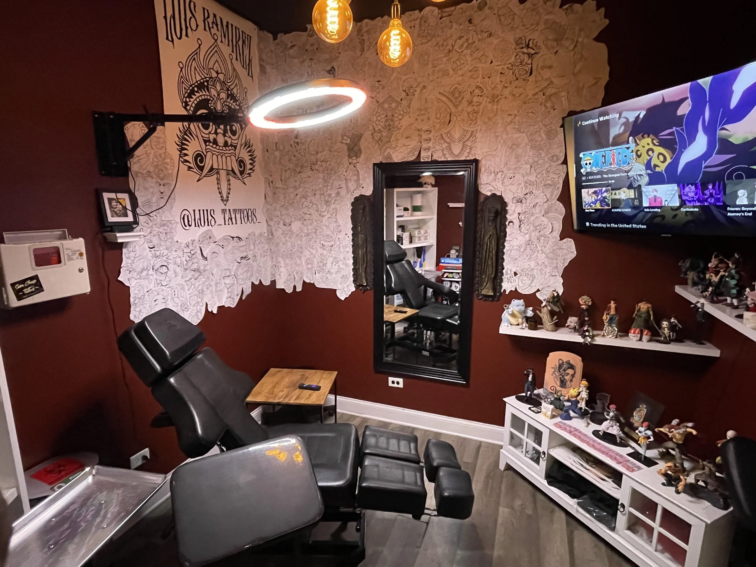 Tattoo studio with a tattoo chair, a large wall mirror, and shelves filled with figurines. The wall features a tattoo artist's logo and a large mural sketch. There is a TV on the wall displaying a streaming service. The room has dim lighting with decorative hanging bulbs.